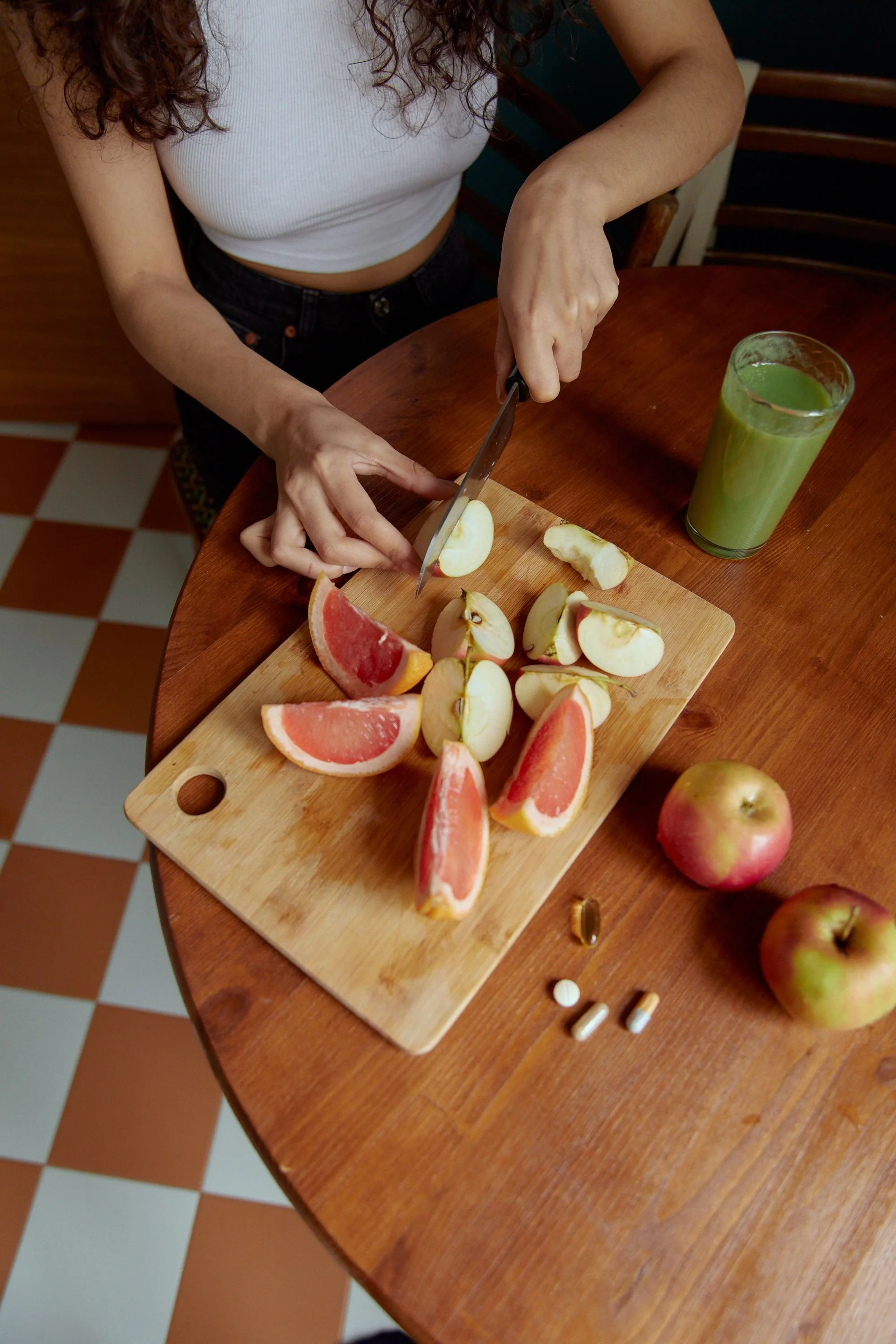 A person is slicing an apple on a wooden cutting board, with grapefruit slices and a green smoothie nearby on a round wooden table. There are also two apples and some pills on the table.