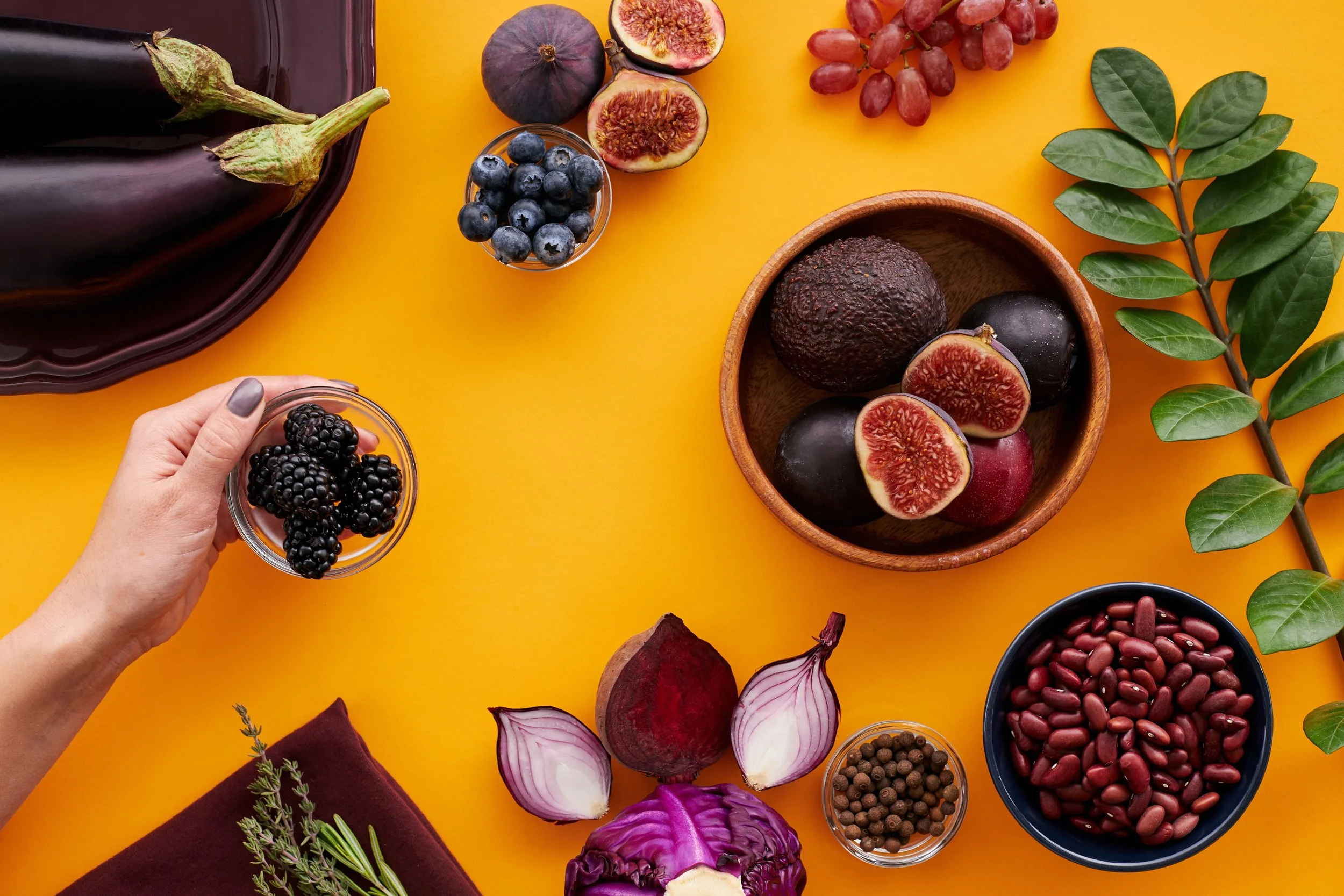 Various fresh fruits including eggplants, figs, blueberries, red grapes, blackberries, and other berries arranged on a yellow background with some vegetables and leaves.