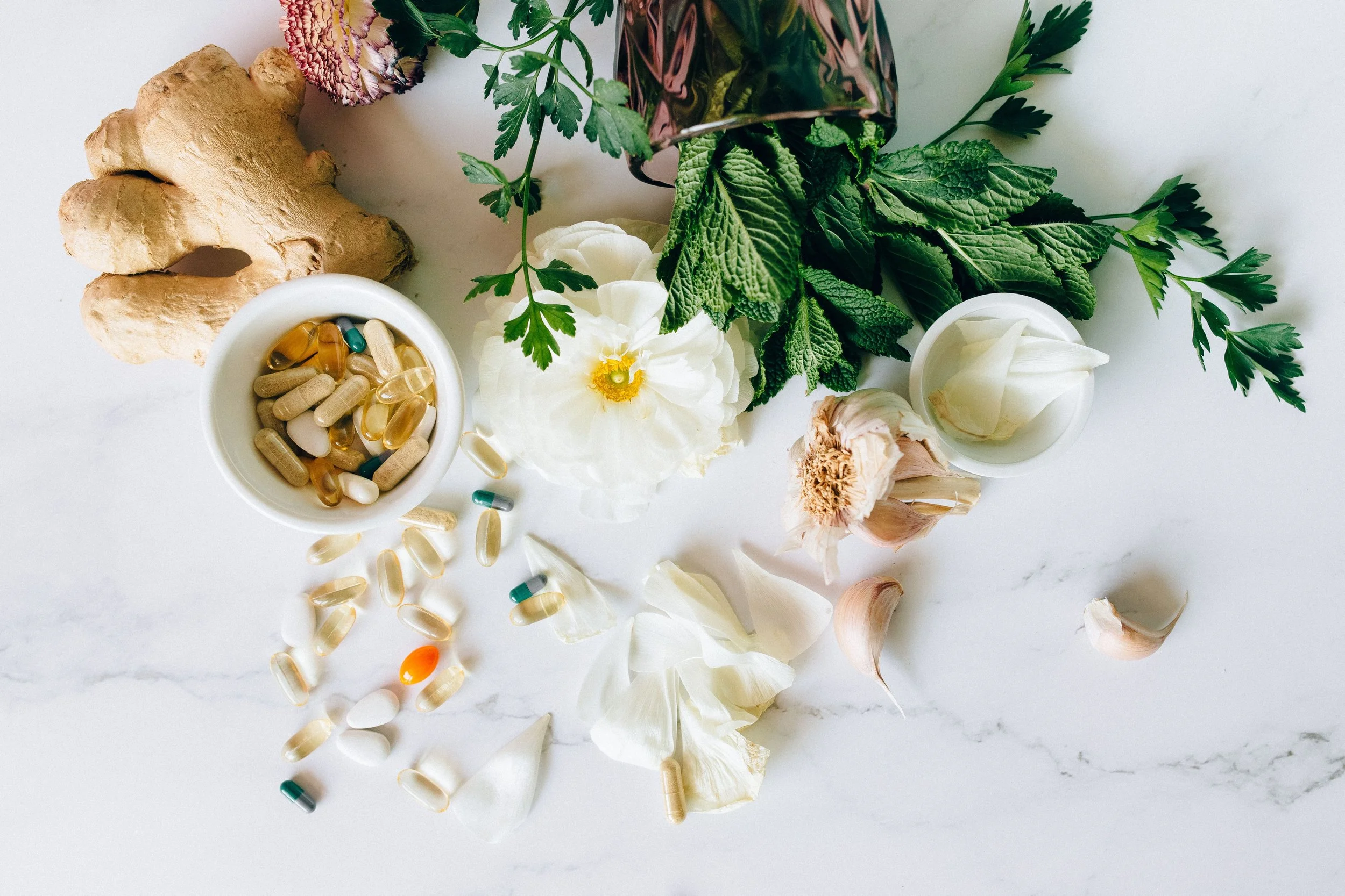 A flat lay of herbs, ginger root, garlic, and various pills and capsules on a white surface.