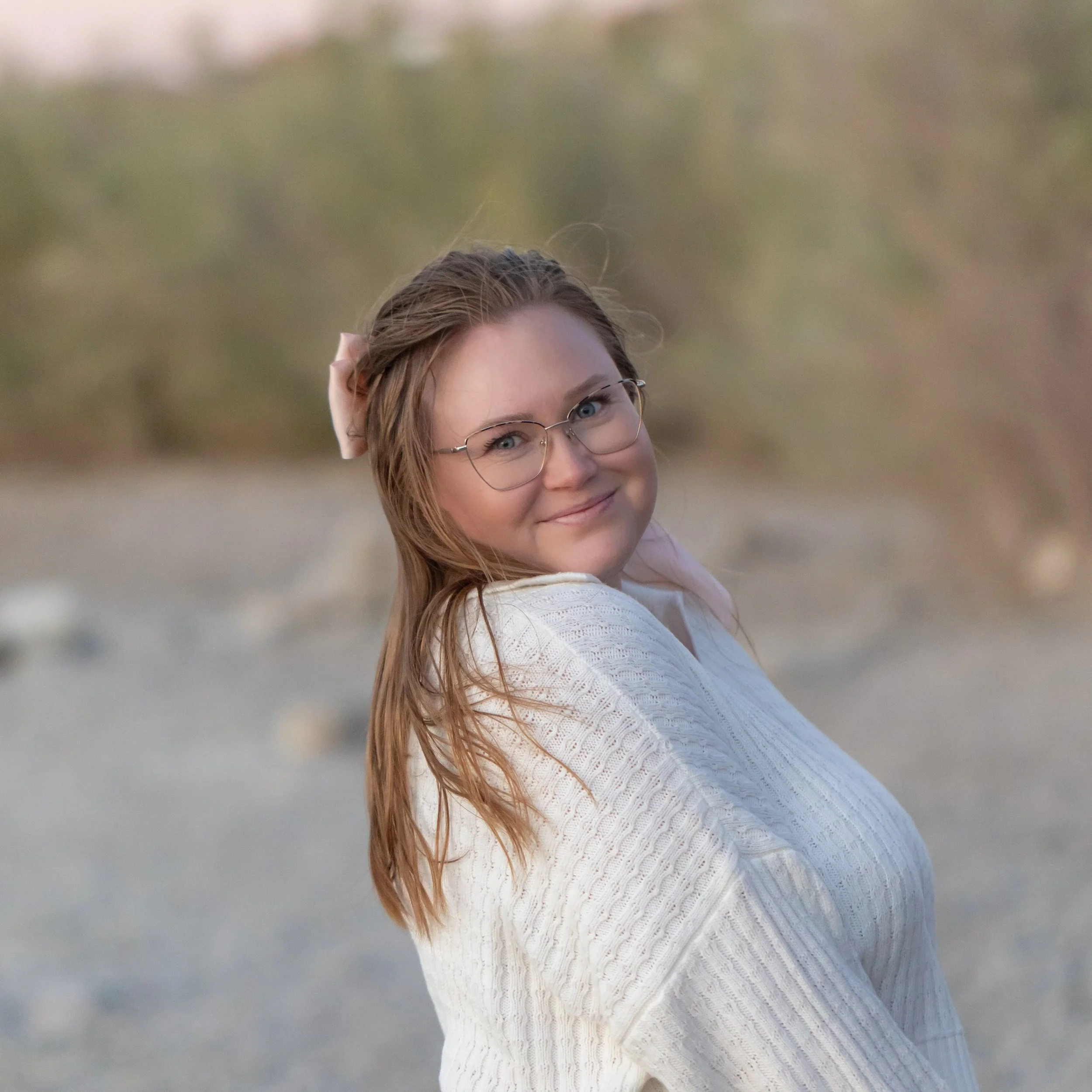 A young woman with glasses and long hair, smiling and wearing a white sweater, standing outdoors with a blurred background of trees and rocks.