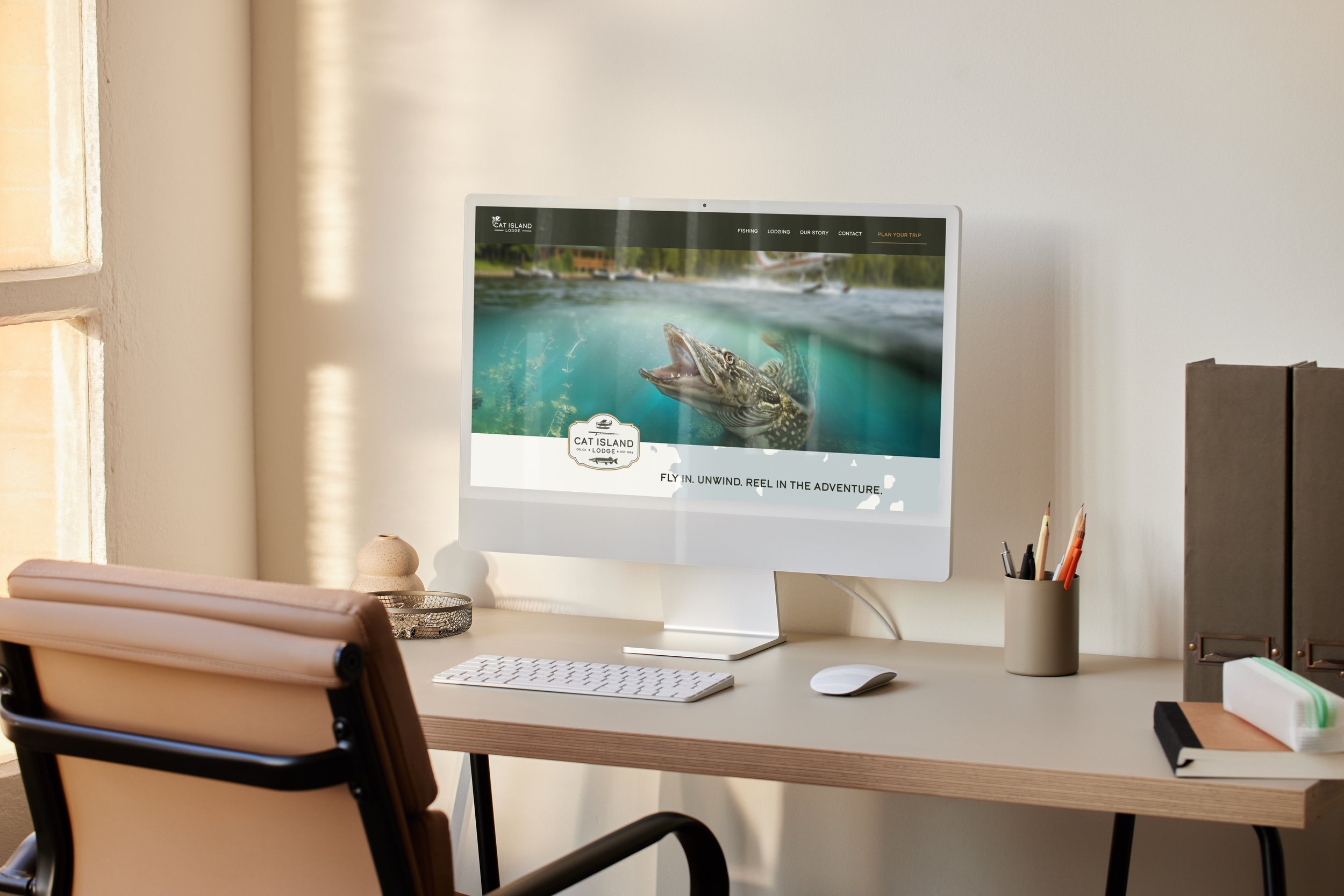 Desk with a computer monitor displaying a website about fishing trips, a keyboard, a mouse, a pen holder, and binders in a well-lit room.