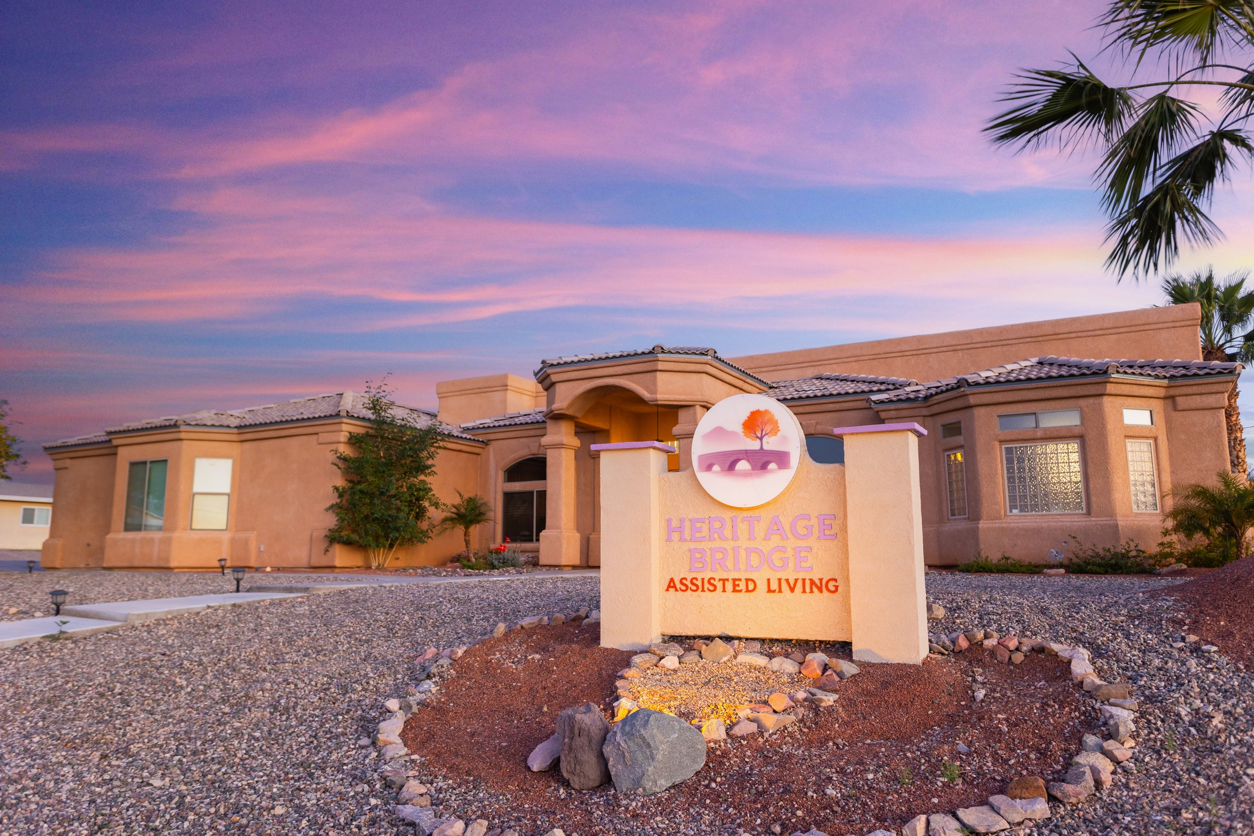A single-story beige building with a sign that reads 'Heritage Bridge Assisted Living' in purple and orange letters, under a pink and purple sunset sky, with desert landscaping and palm trees.