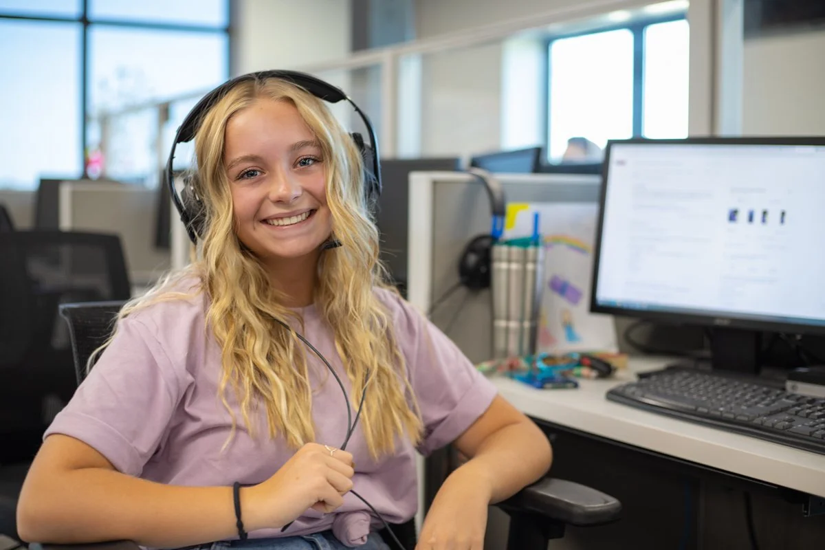 A young woman with long blonde hair smiling at the camera while wearing headphones in an office environment, with computer monitors and work supplies on her desk.
