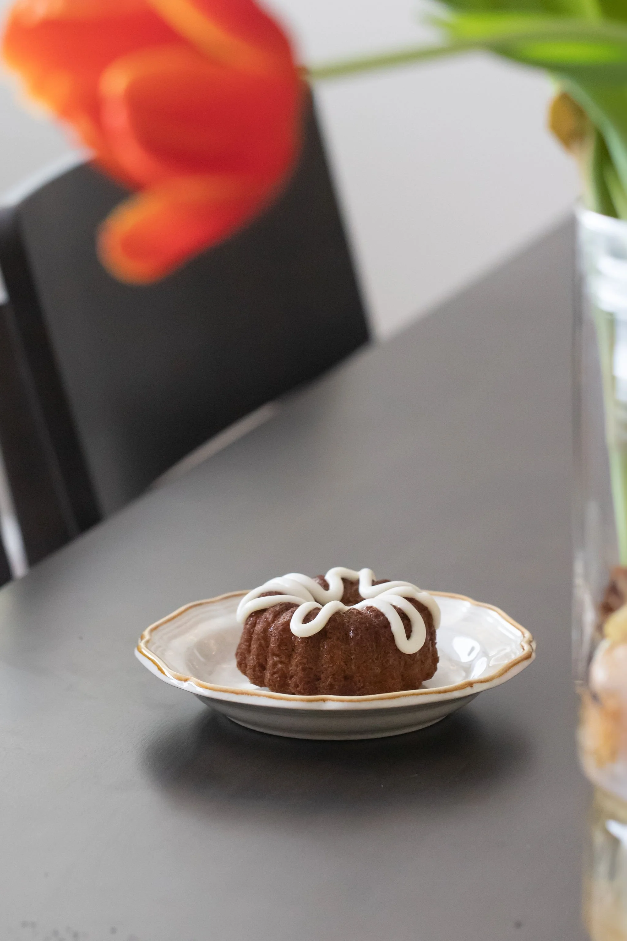 Chocolate bundt cake with white icing on a decorative white plate with gold trim, on a gray table.