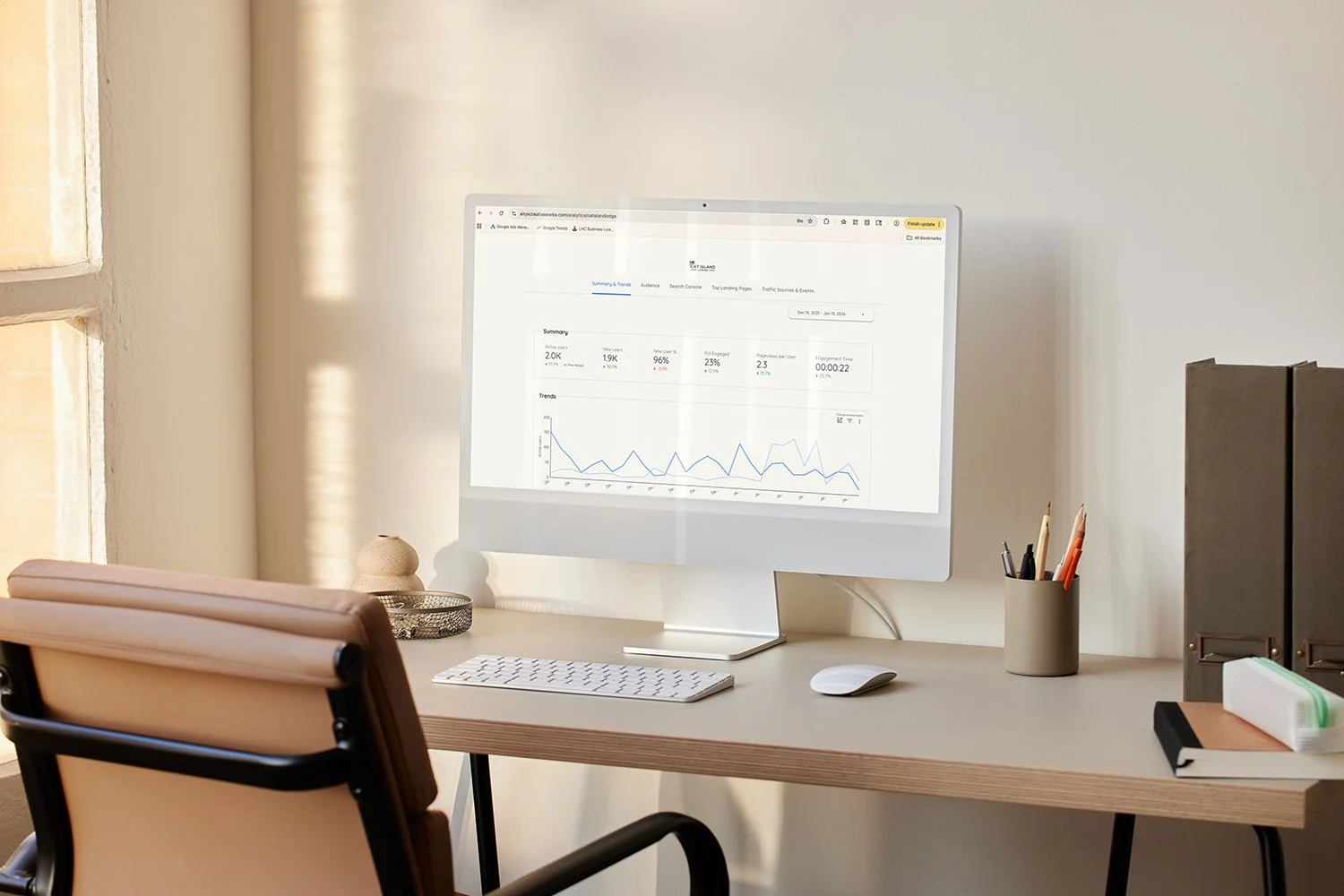 An organized home office desk with a large computer monitor displaying analytics, a wireless keyboard, a mouse, a pencil holder with pens and pencils, a beige chair, and a window letting in natural light.