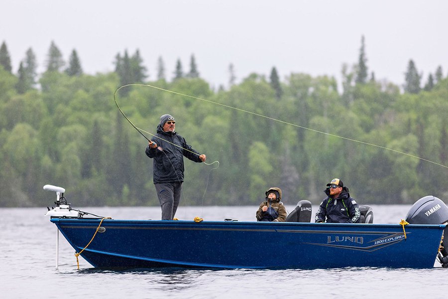 Three people fishing on a blue boat on a lake with a forest in the background.
