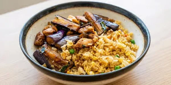 A bowl of fried rice with cooked egg, green peas, and eggplant slices on the side.