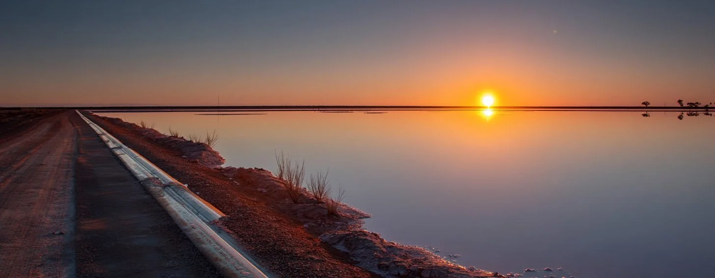 Sunset over a calm body of water, with a dirt road running alongside it and some sparse vegetation.