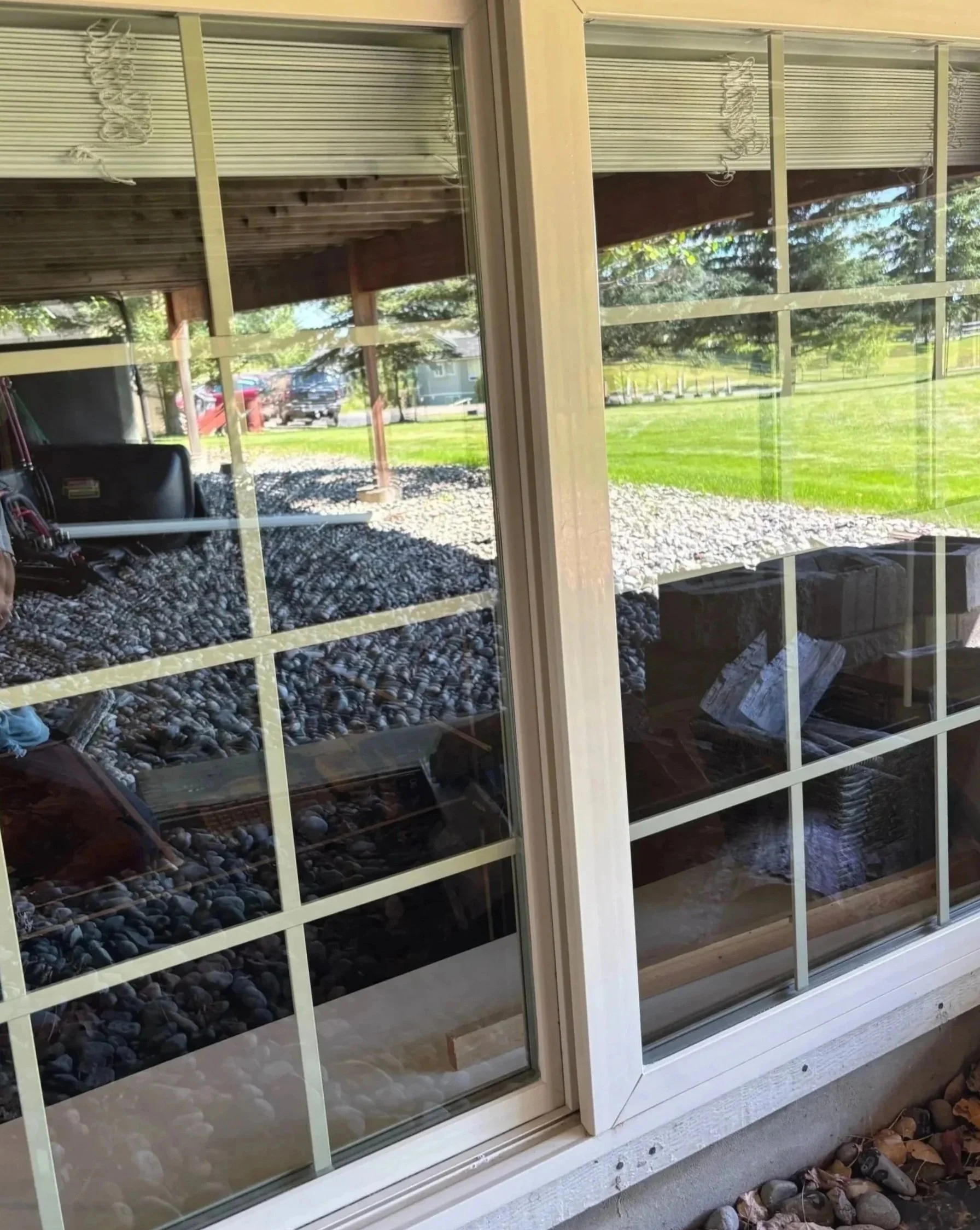 Close-up view of a sliding glass door with a white frame, showing reflections of trees and a grassy yard outside, with some rocks on the ground outside.