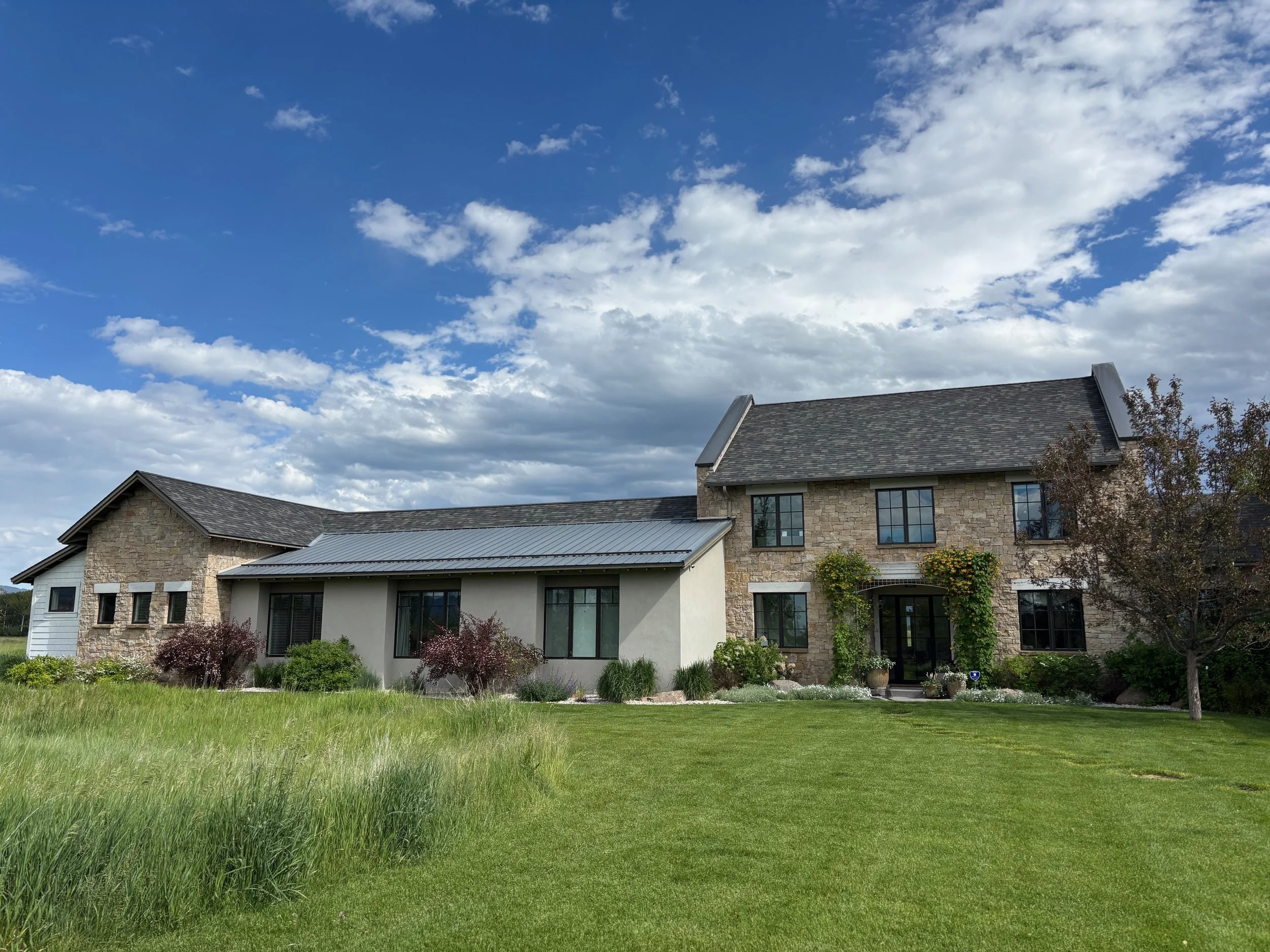 A large house with stone and white exterior walls, multiple dark windows, and a gray roof, set against a partly cloudy sky with green landscaping and trees in the front yard.