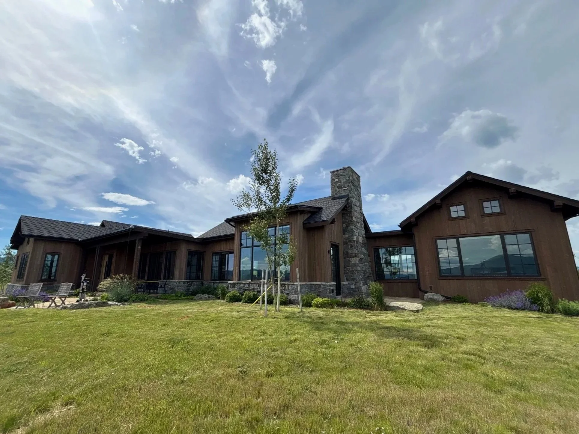 Modern house with large glass windows, a chimney, and wooden exterior, surrounded by a lawn, small trees, and outdoor seating, under a partly cloudy sky.
