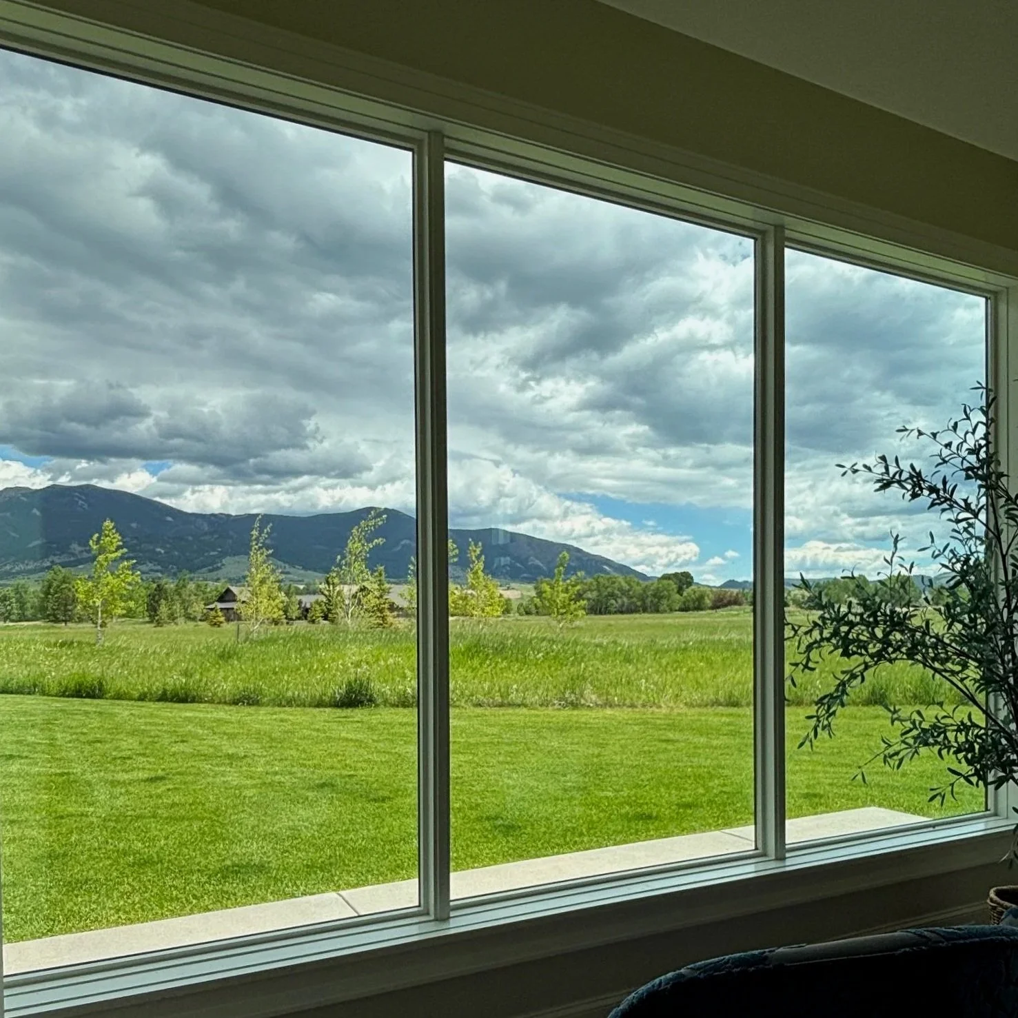 View through a large window showing green grass, trees, and mountains under a cloudy sky.