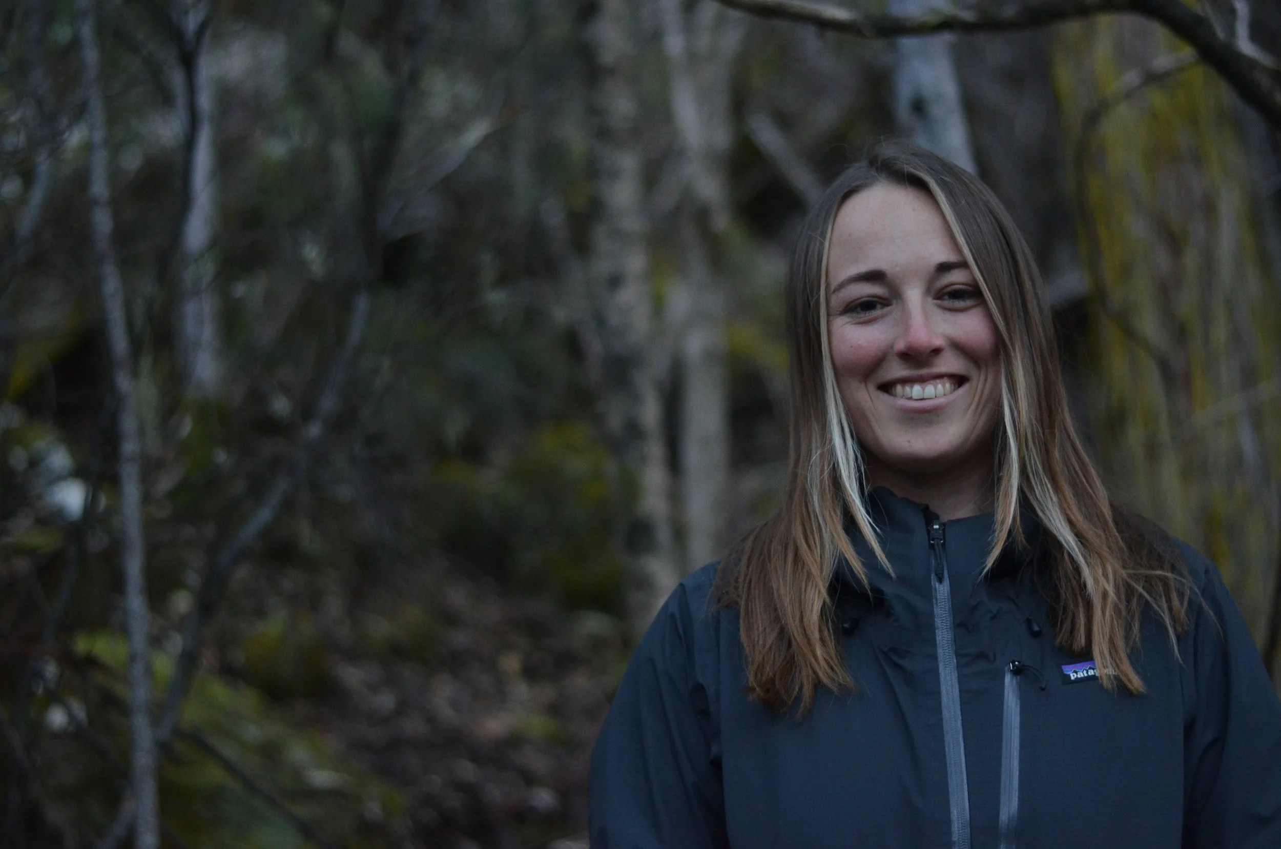 A woman with long blonde hair smiling outdoors surrounded by trees and foliage.