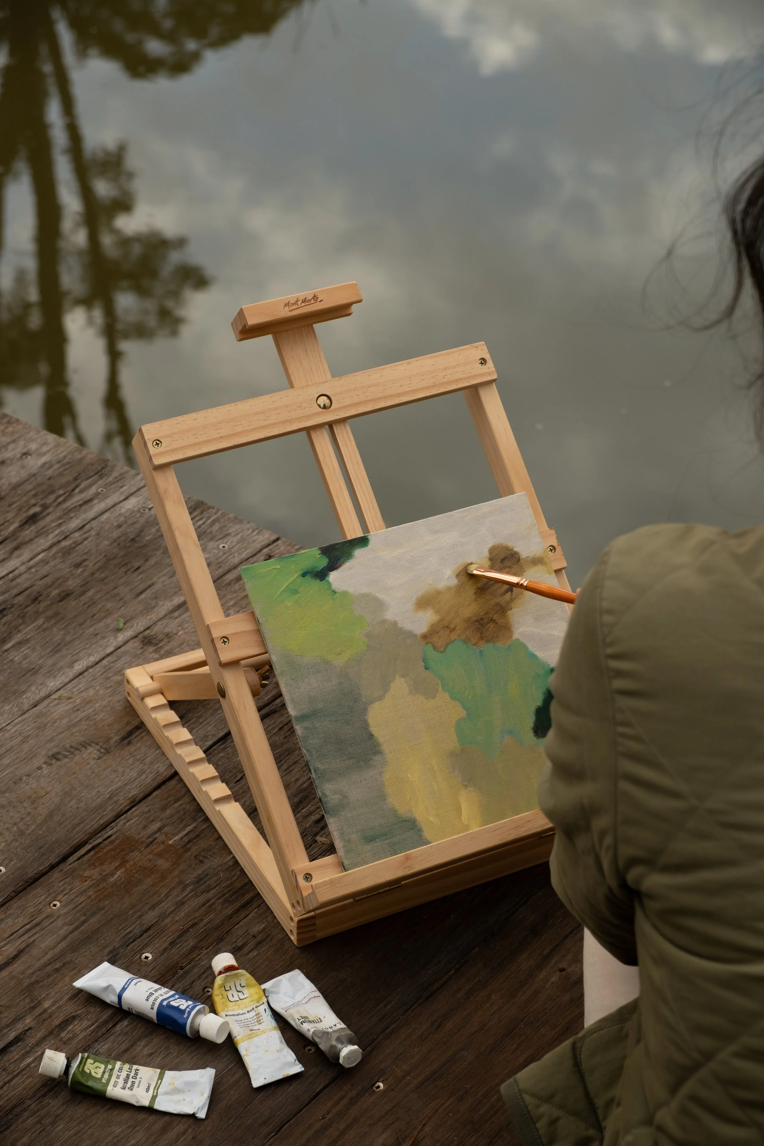 A person painting a landscape using oil paints on a canvas, set on a small wooden easel by a body of water. Several tubes of paint are laid out on a wooden surface nearby.