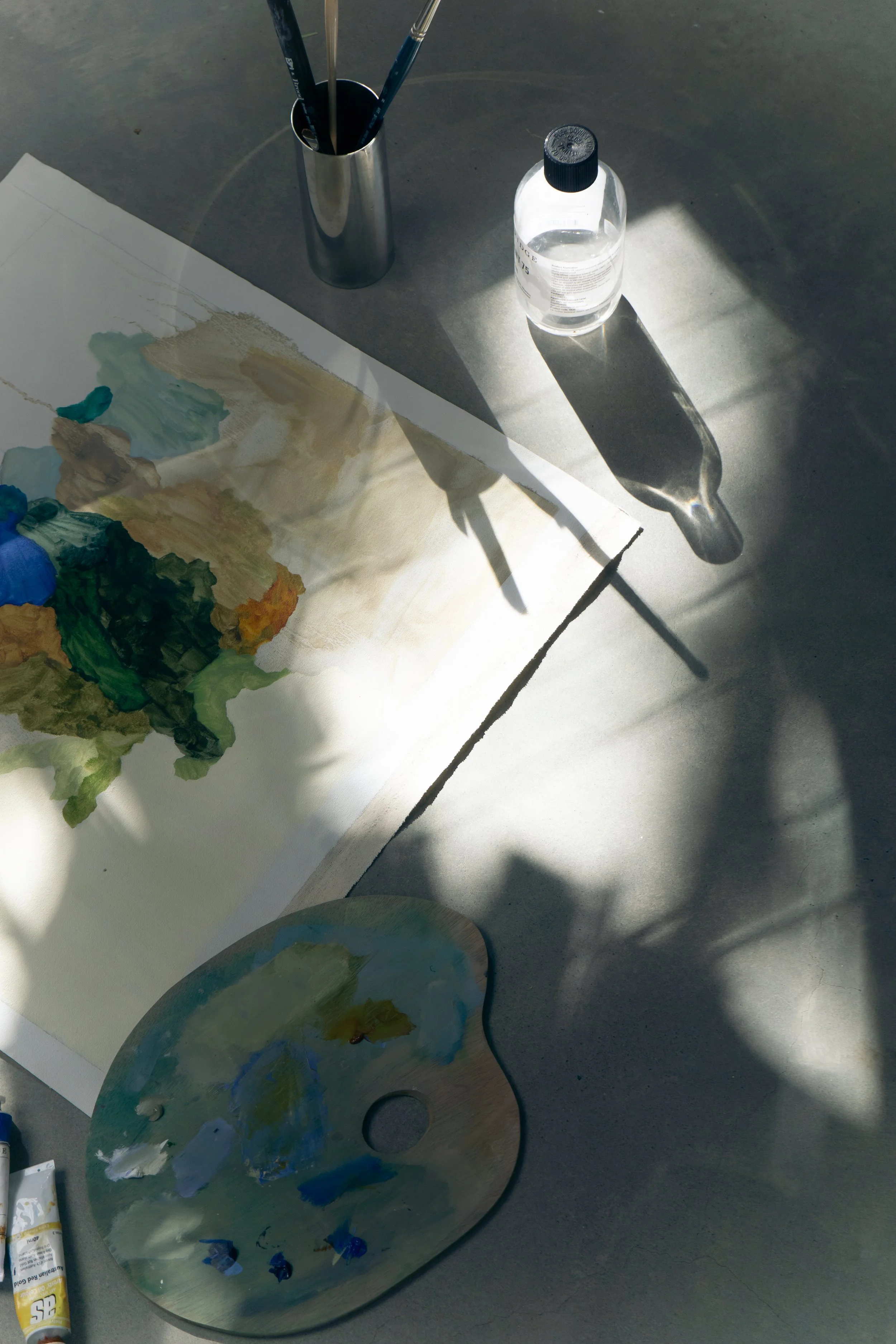 A still life scene of an artist's workspace featuring a paint palette with mixed colors, a partially painted artwork on paper, a small palette with paint, a metallic container with brushes, and a tube of yellow paint, all on a gray surface with shadows from sunlight.