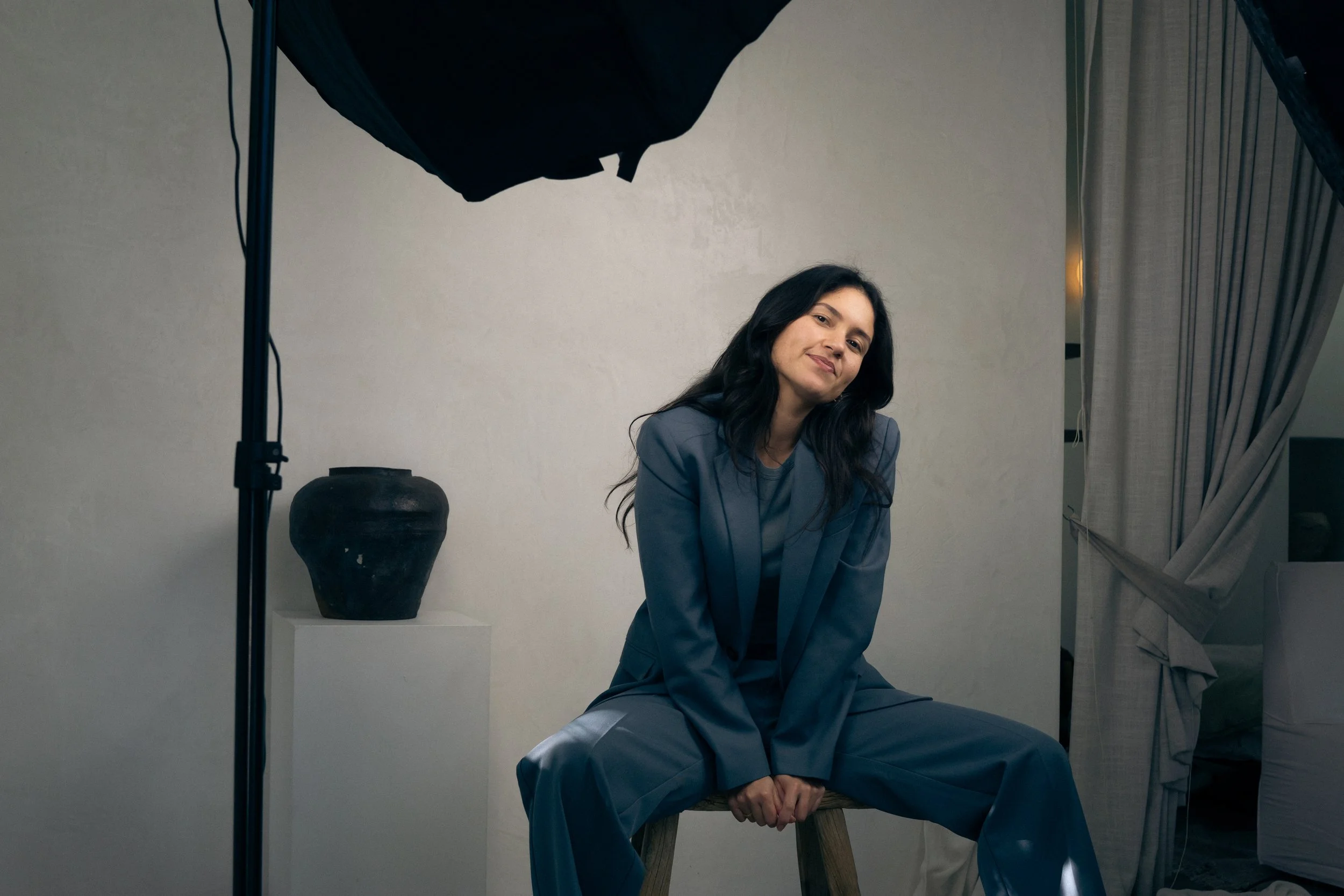 A woman with long dark hair, wearing a St Agni suit, sitting on a wooden chair. She is in a photography studio setup with a gray background, a large black reflector or softbox above, and a tall clay pot in the background.