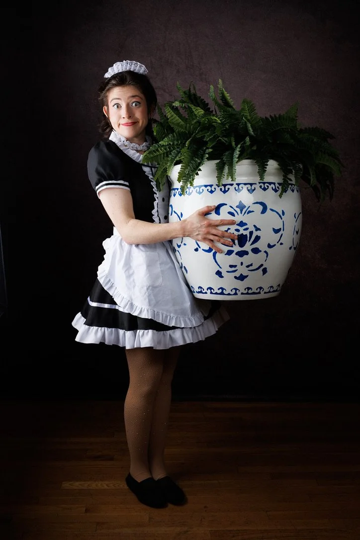 A woman dressed as a maid holding a large potted fern plant with a dark background and wooden floor.