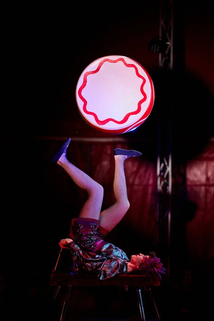 A performer foot juggling a drum at Circus Bella. The performer is wearing fishnet stockings, a shiny sequined jacket, and a feathered headpiece, set against a dark background.