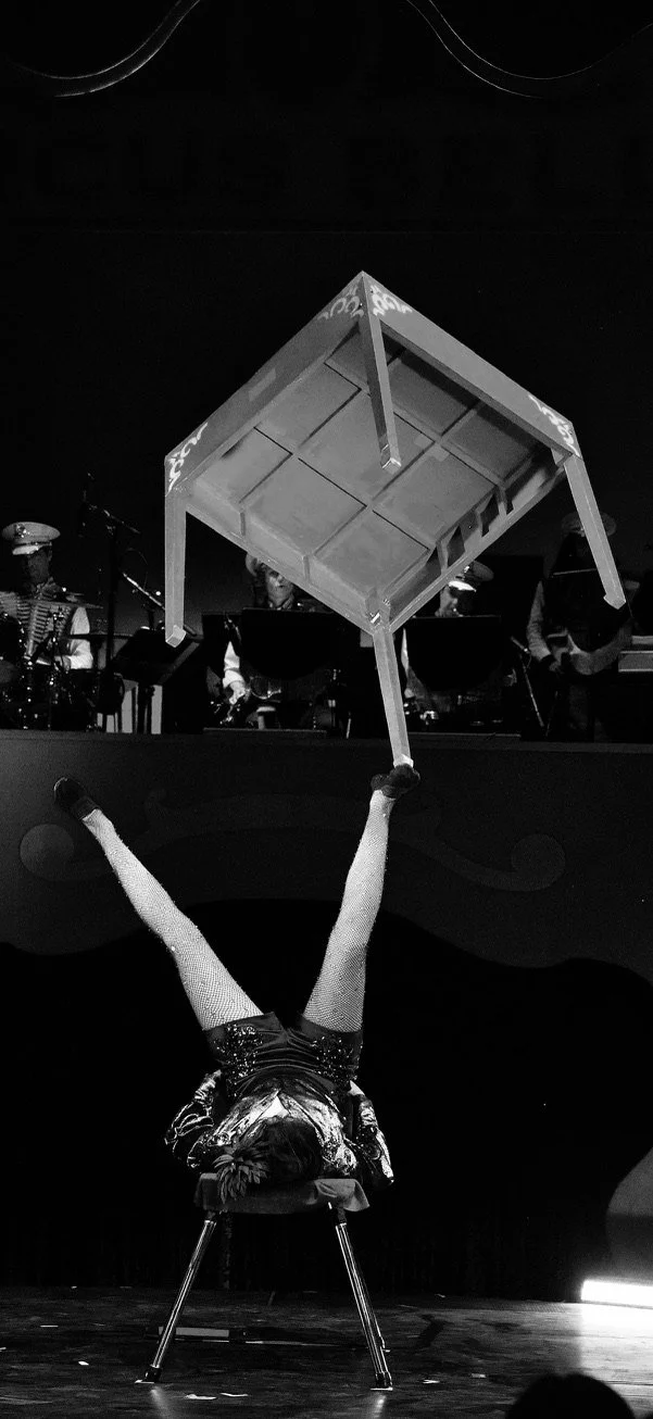 A performer foot juggling a table. Balanced on the feet, with a band playing in the background.