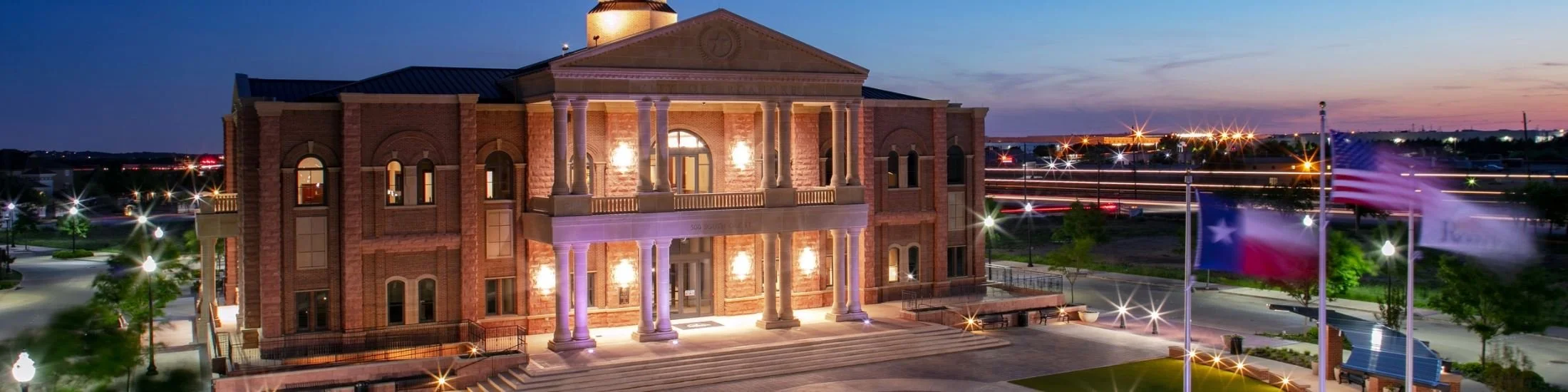 A historic courthouse at dusk illuminated by exterior lights, with a flagpole and American flags in the foreground, and a sunset sky in the background.
