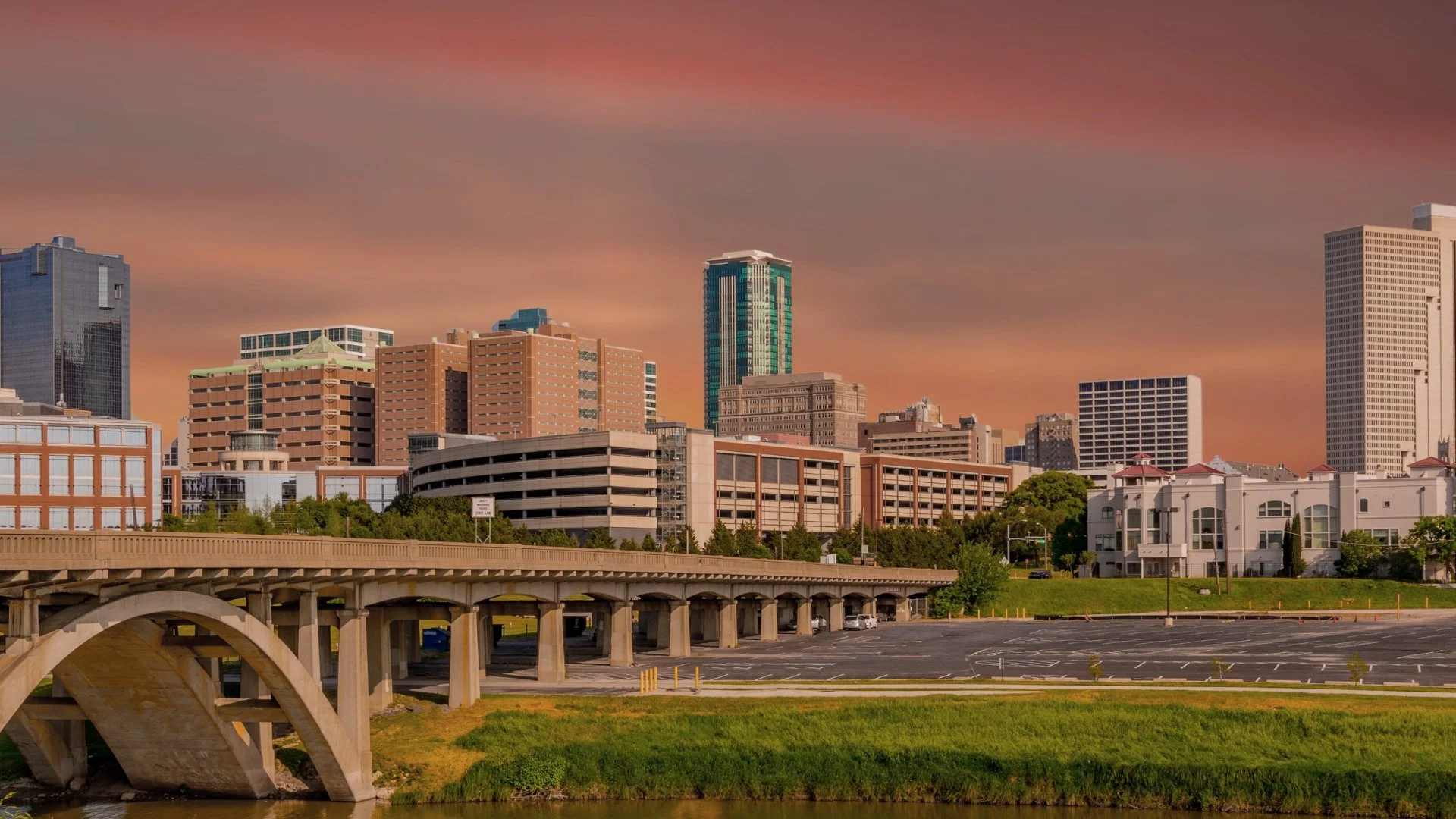 Urban city skyline at sunset with various skyscrapers and buildings, a bridge in the foreground, parking lot, and a grassy area with trees.