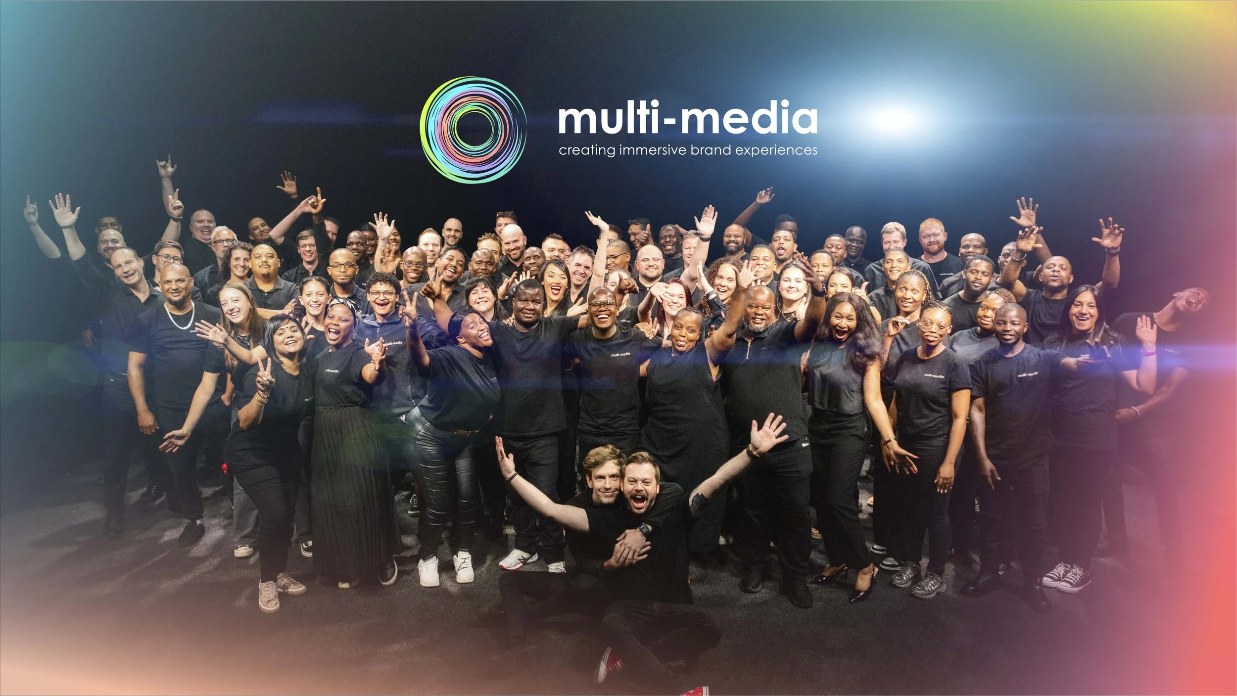 A large group of diverse people smiling and celebrating, posing for a photo in front of a dark wall with the logo and text 'multi-media creating immersive brand experiences'.