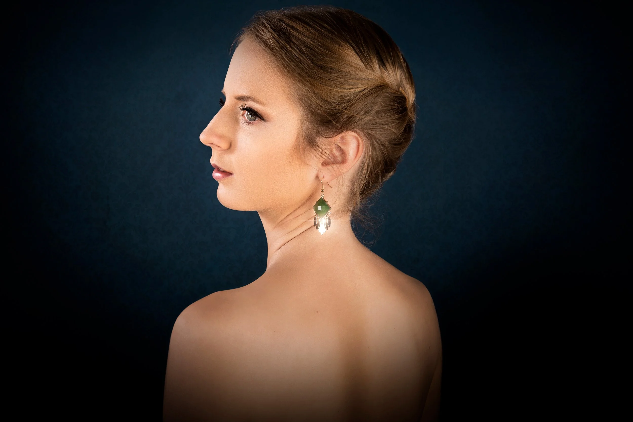 Portrait d'une femme avec cheveux attachés, portant des boucles d'oreilles tombantes vert foncé et argentées, la photo est prise en profil avec un fond sombre.