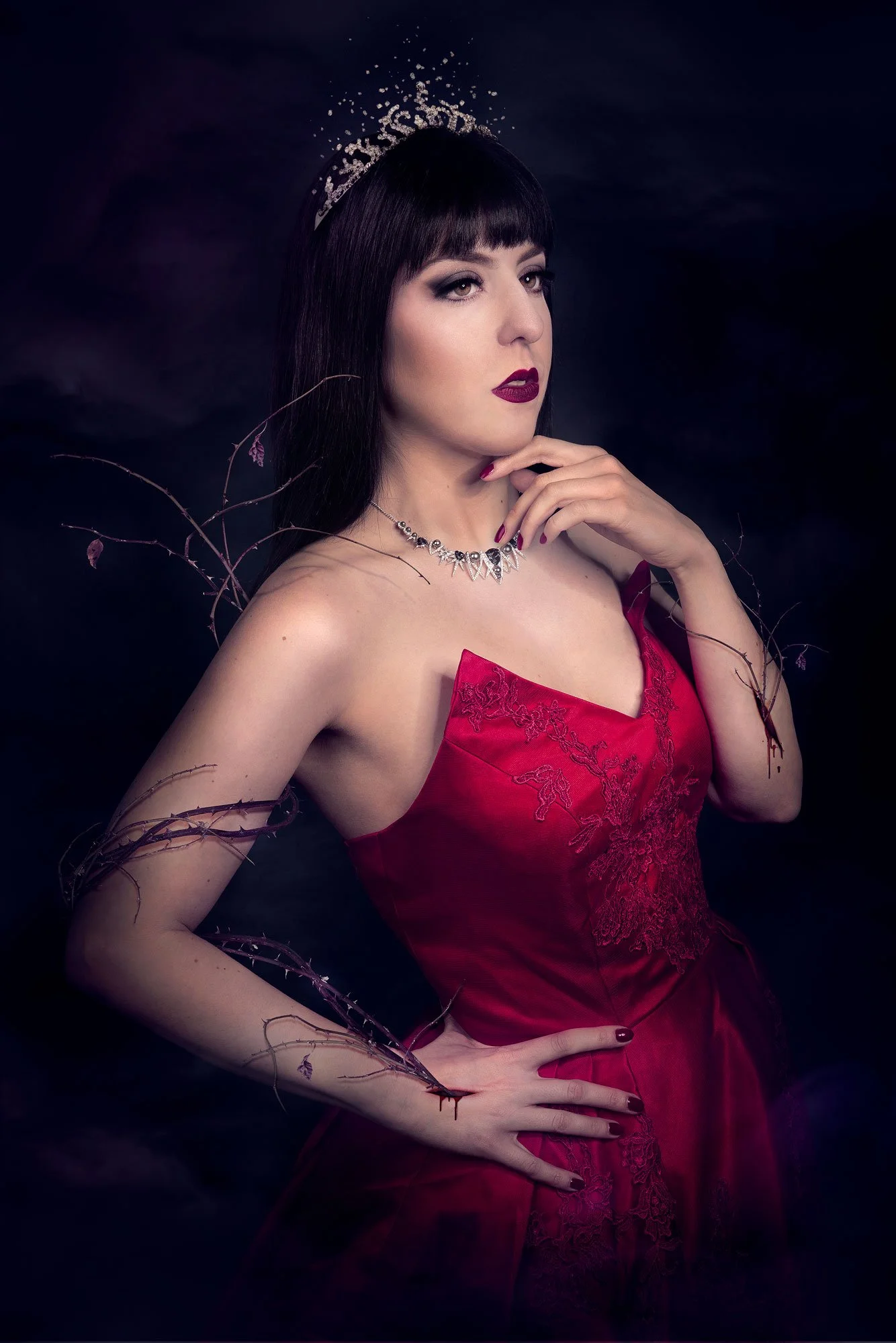 Une femme portant une robe rouge, un collier élégant, et un maquillage sombre, avec une couronne de strass, pose avec confiance dans un fond sombre, entourée de branches sèches.