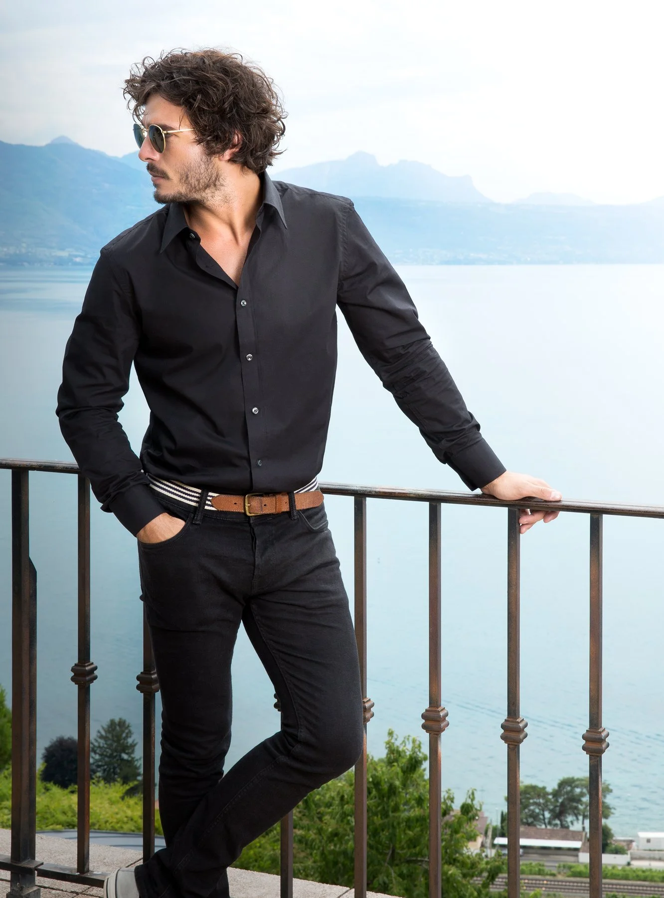 Homme portant des lunettes de soleil, en chemise noire, debout devant un lac avec des montagnes en arrière-plan, tenant une rambarde.