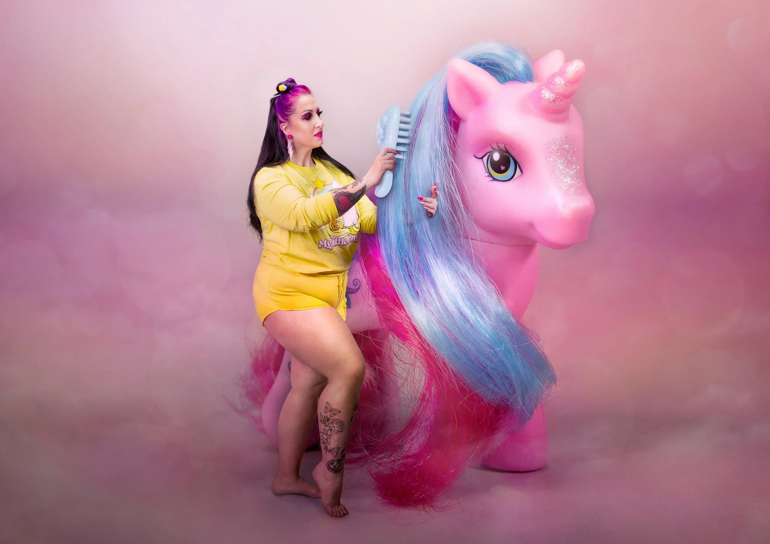 Une femme avec des cheveux roses et violets se brosse les cheveux devant un grand masque de licorne rose avec une corne scintillante et des yeux grands et colorés, dans un décor rose pastel.