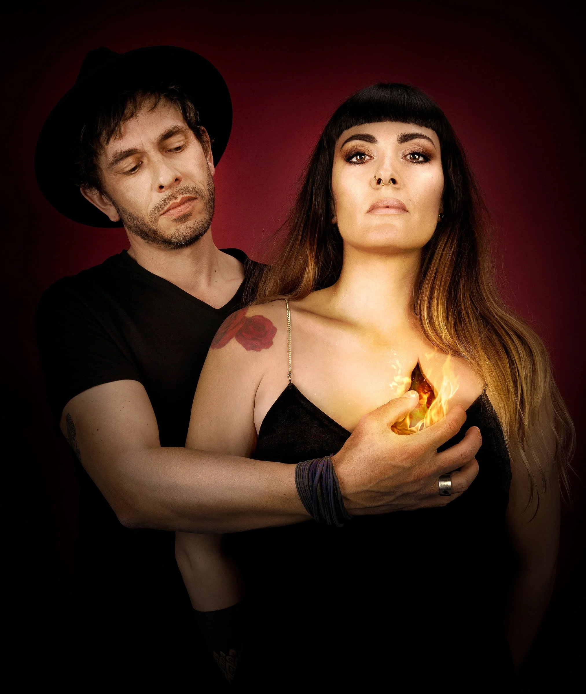 Une femme avec un tatouage de rose sur l'épaule tient une boule de feu dans la paume de sa main, entourée d'une ambiance sombre avec un fond rouge foncé. Un homme la regarde avec sérieux.