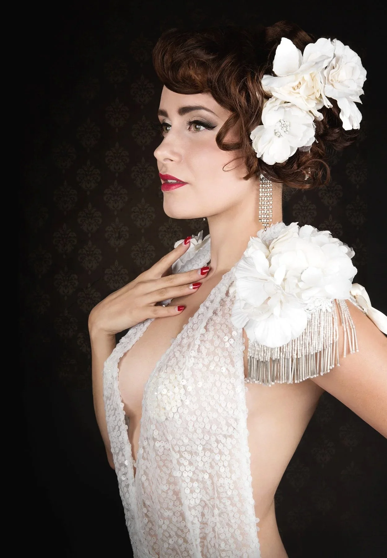 Une femme élégante avec des cheveux bouclés ornés de grandes fleurs blanches, portant de longues boucles d'oreilles et une robe en dentelle blanche avec des détails argentés, pose en profil devant un fond noir décoratif.