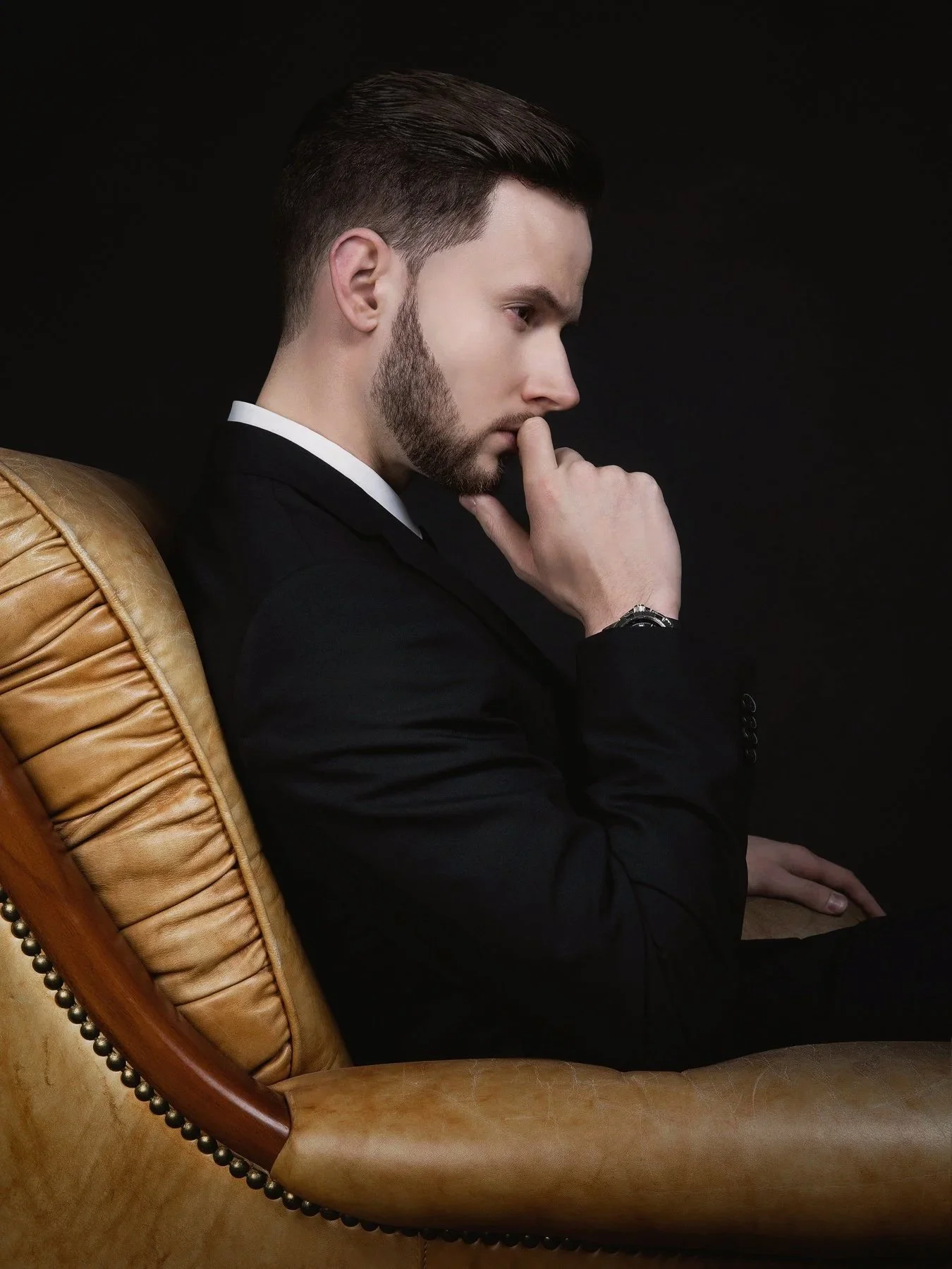 Un homme en costume noir est assis sur un fauteuil en cuir beige, avec un fond noir. Il a les cheveux courts, une barbe et tient sa main près de sa bouche dans une posture pensive.