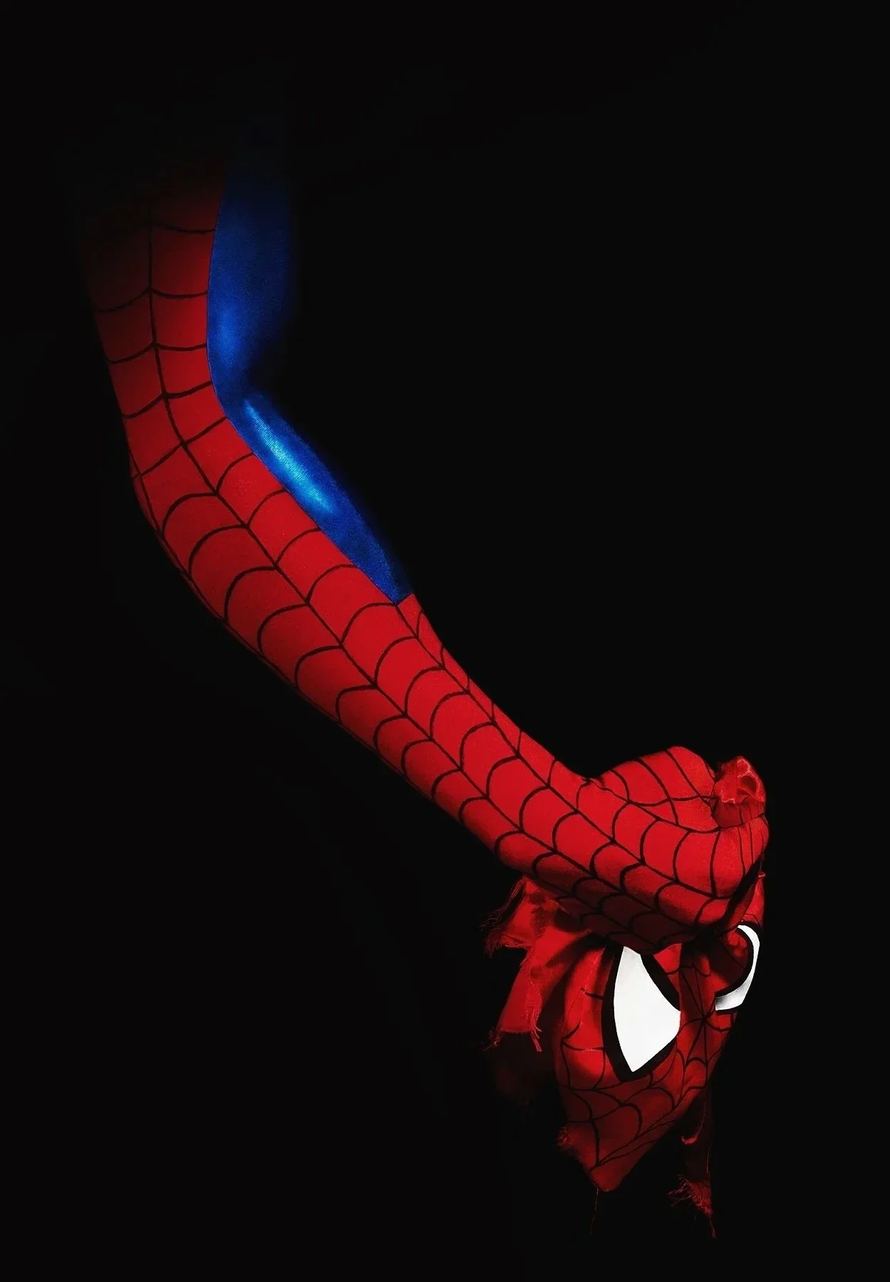 Une personne portant un costume de Spider-Man, avec un masque rouge et des yeux blancs, soulève la manche de son costume pour révéler la partie bleue et rouge de la tenue, tout en étant dans un fond noir.