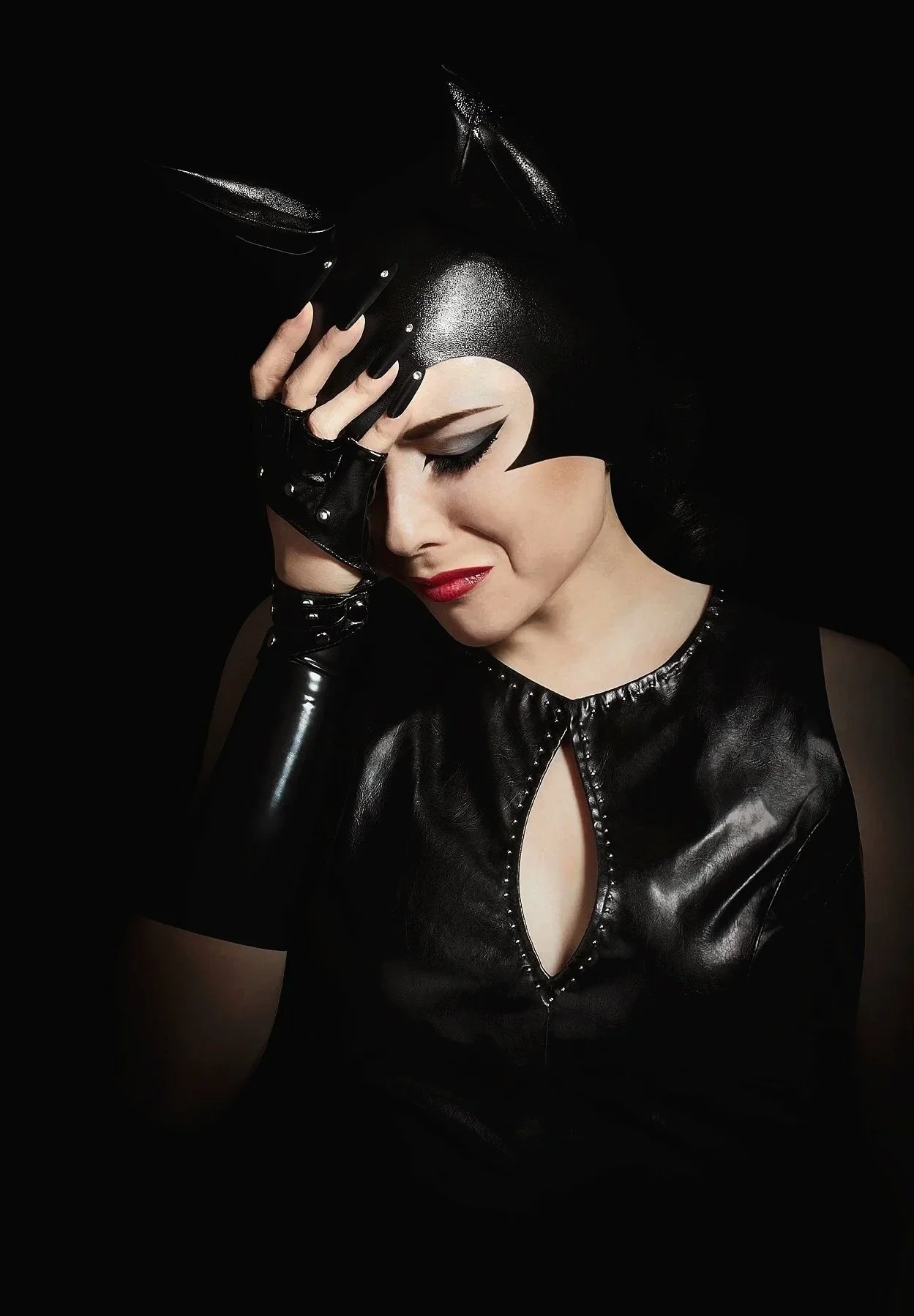 Une femme porte une tenue en cuir noir avec des gants, ses yeux sont maquillés avec du maquillage noir et ses lèvres sont rouges. Elle se tient contre un fond noir.