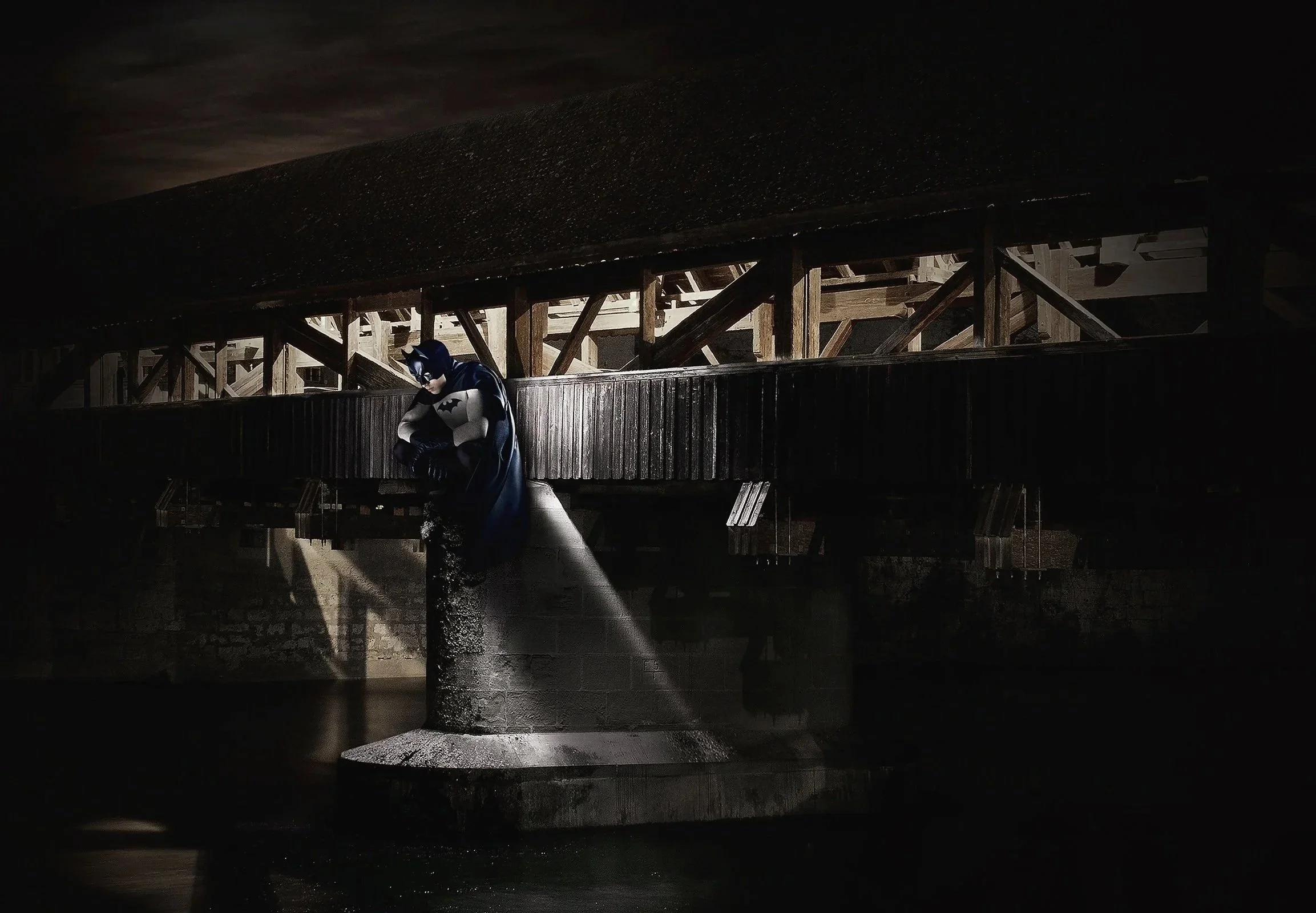 Un homme déguisé en Batman, debout sous une fontaine la nuit, avec un environnement sombre et une structure en bois en arrière-plan.