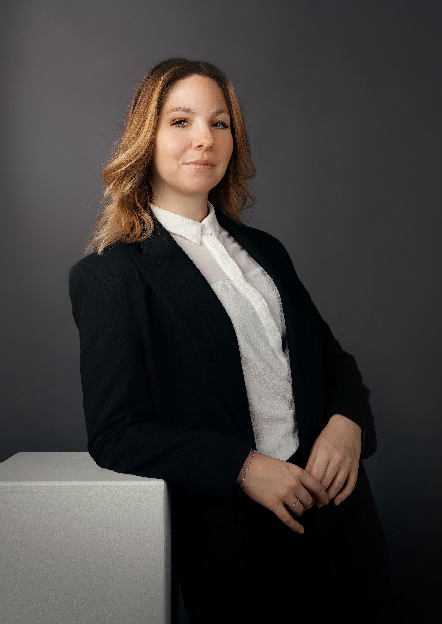 Femme d'affaires en costume noir avec une chemise blanche, posture confiante, fond gris foncé.