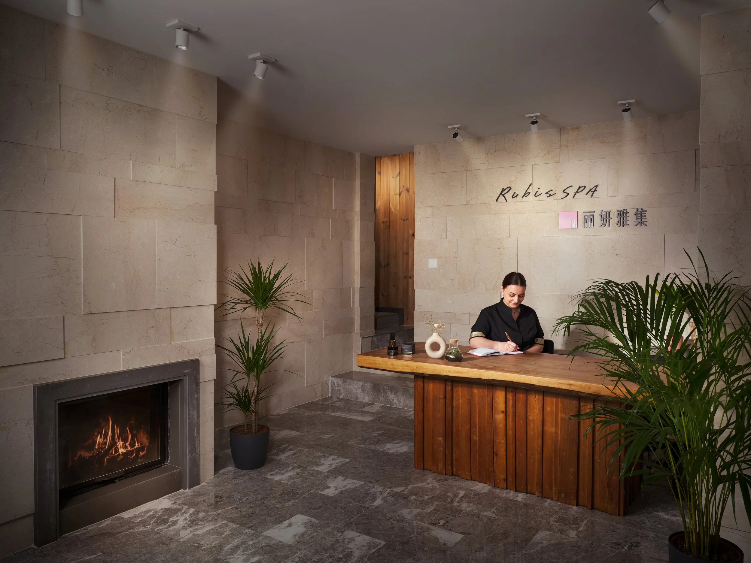Intérieur d'un spa ou salon de massage avec un mur en pierre, un foyer au bois allumé, un comptoir en bois avec une femme qui écrit dans un carnet, des plantes vertes, et un signe indiquant 'Rubis SPA'.