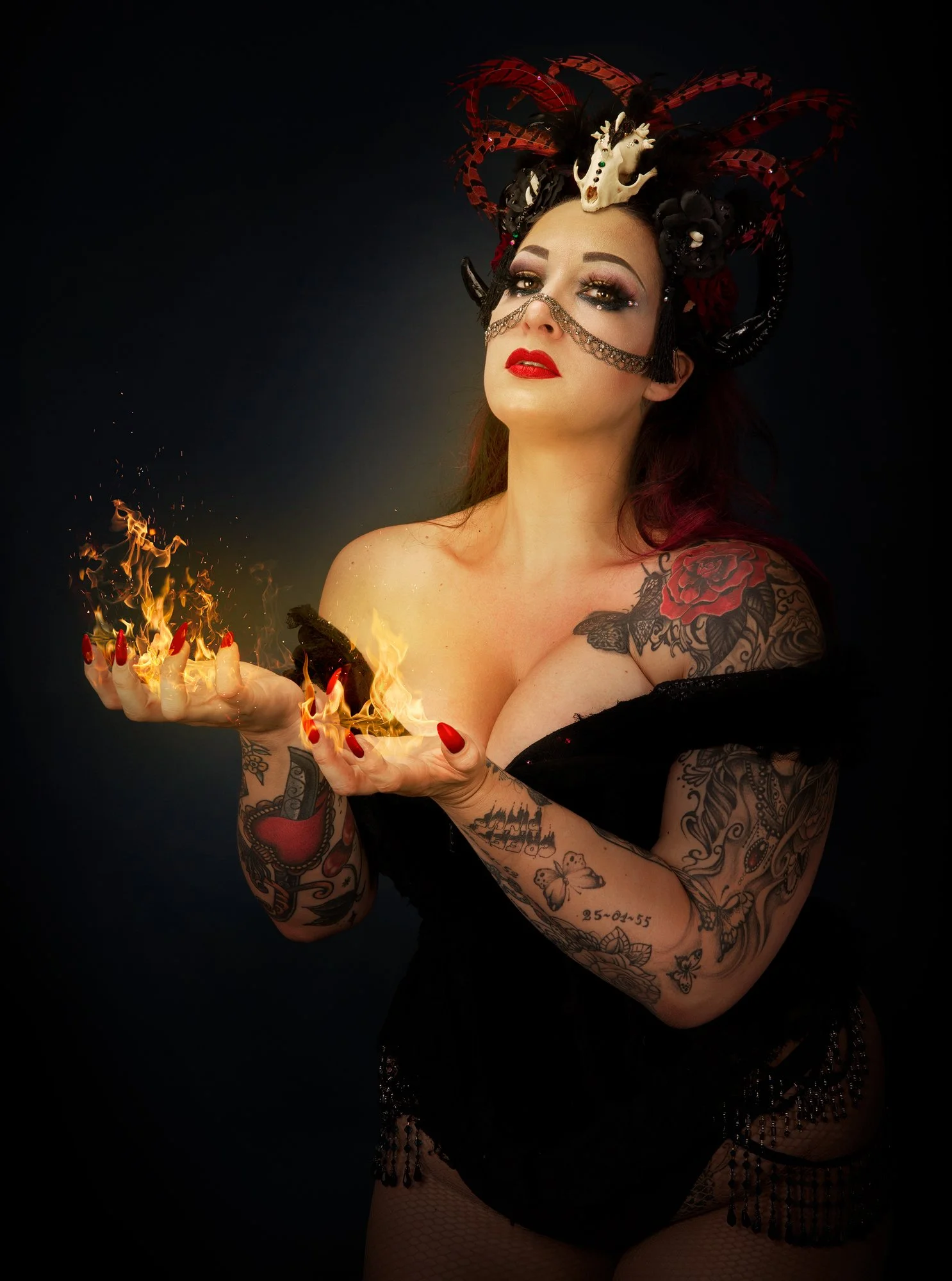 Une femme tatouée portant un déguisement sombre et orné de plumes et d'un crâne sur la tête, tenant un gâteau en feu dans la main.