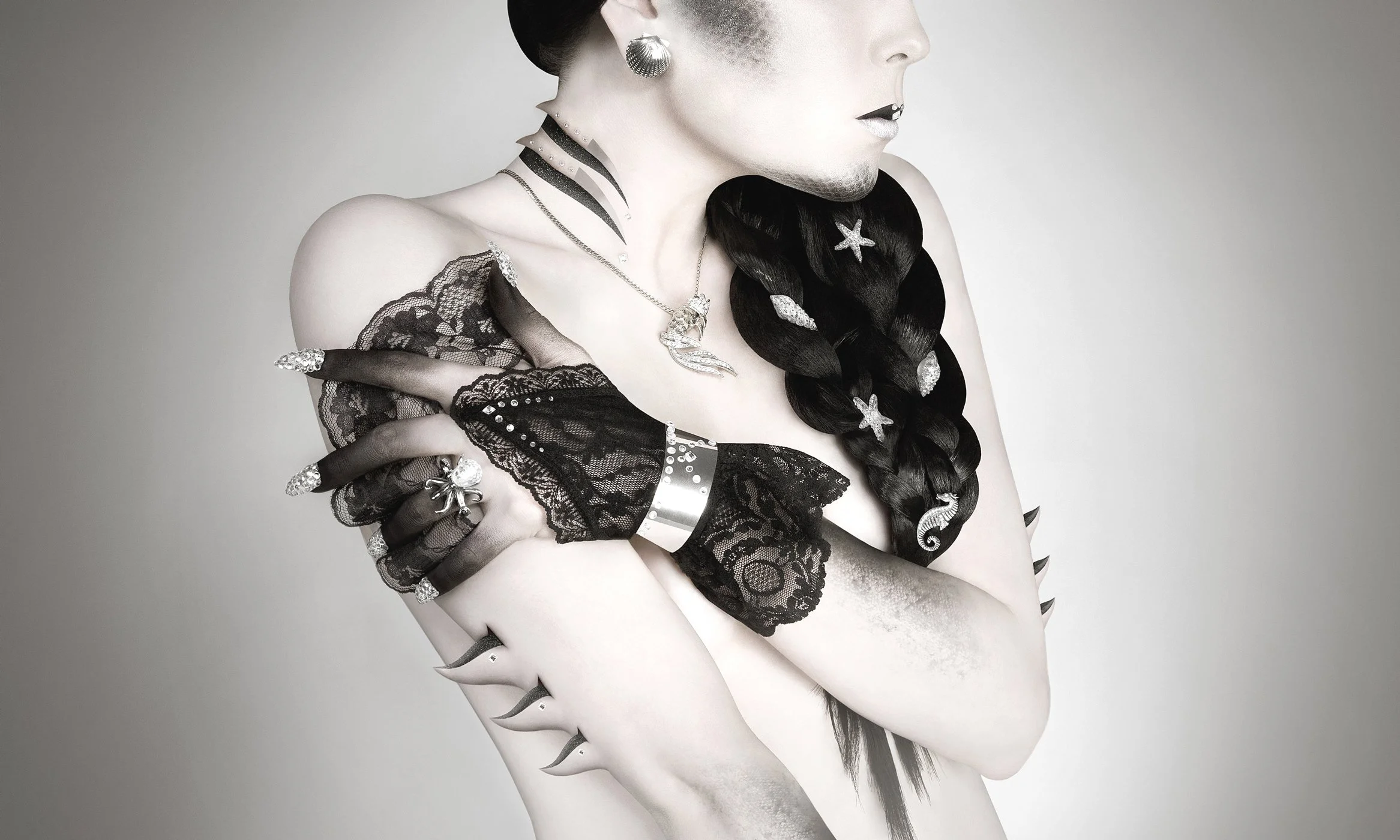 Une femme au profil, portant des accessoires en bijoux en argent avec des détails en strass, des vêtements en dentelle noire, avec une coiffure tressée ornée de décorations marines telles qu'une étoile de mer et une hippocampe, en noir et blanc.