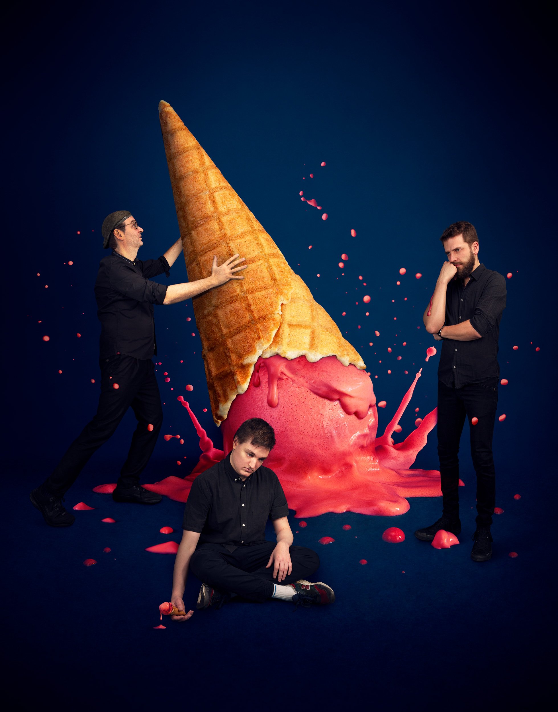 Quatre hommes noirs et blancs posent avec une grande glace en forme de cornet de crème glacée, entourés de éclaboussures de peinture rose. Deux hommes sont debout, dont un qui pousse la glace, et deux autres, assis ou à genoux, sont en position de ré