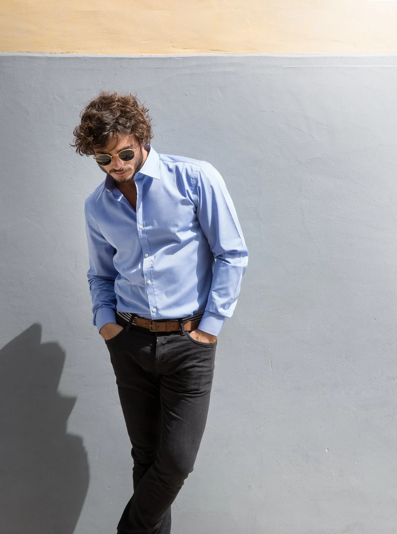 Jeune homme portant une chemise bleue, des lunettes de soleil, avec les mains dans les poches, posant contre un mur gris avec une bande beige en haut.