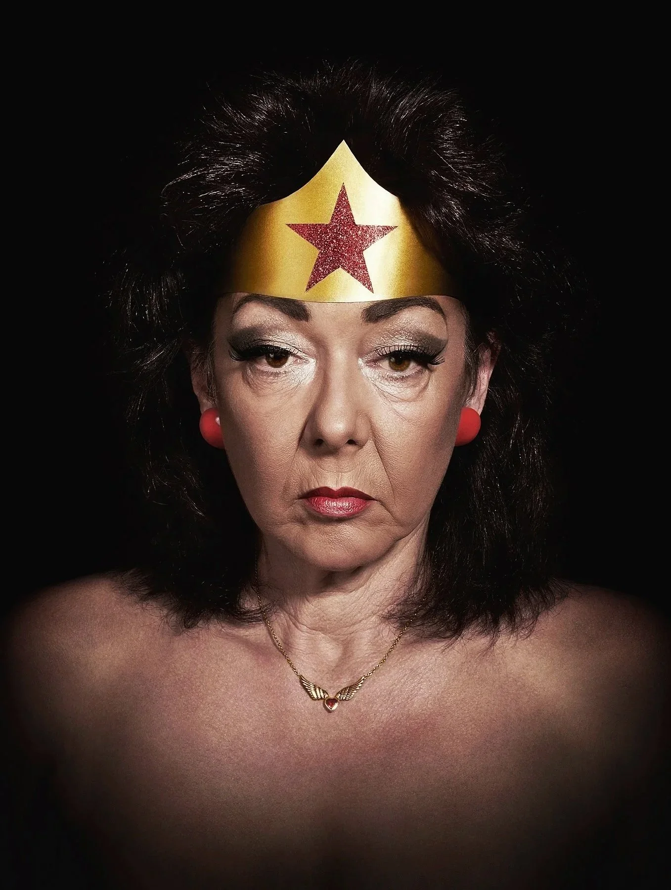 Portrait d'une femme avec un bandeau de Wonder Woman, un maquillage prononcé, un collier avec un cœur et des boucles d'oreilles rouges, contre un fond noir.