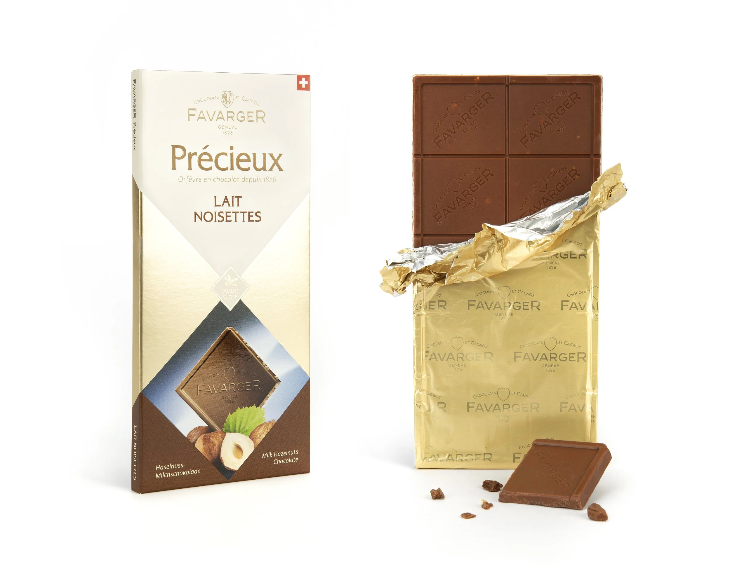 Tablette de chocolat au lait aux noisettes et boîte de chocolat Favarger. La tablette est partiellement déballée, avec quelques morceaux cassés et des éclats autour.
