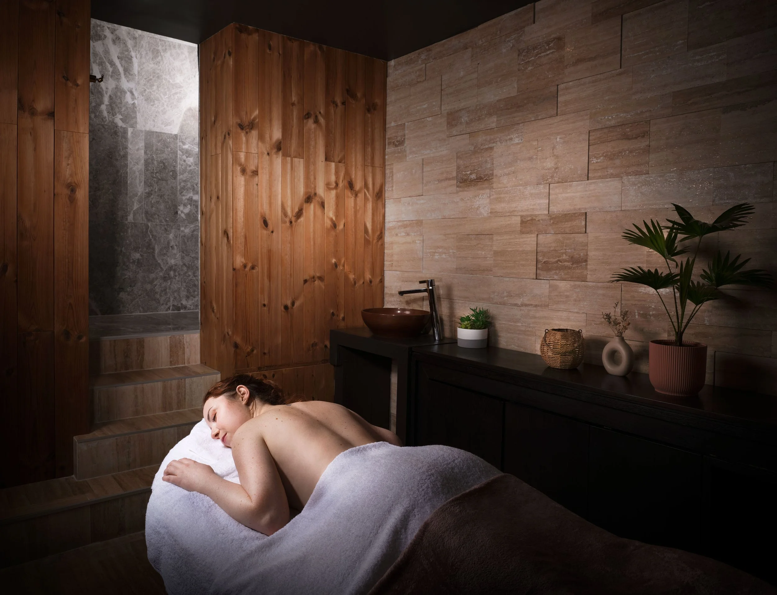 Une femme allongée sur une table de massage dans un spa ou un salon de massage, entourée de plantes et de décorations en bois, avec un mur en pierre derrière elle.