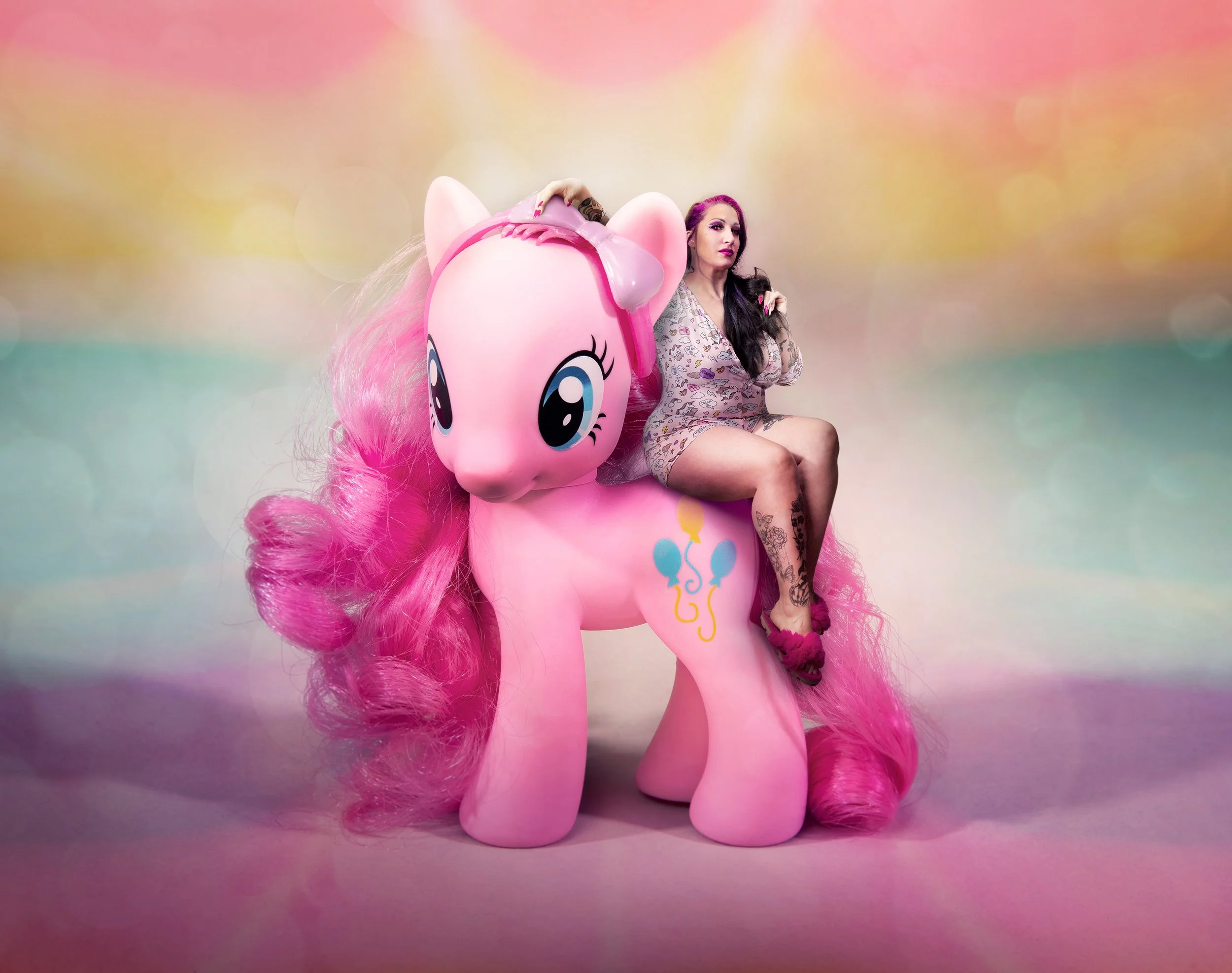 Une femme aux cheveux noirs et violet, avec un maquillage foncé, assise sur une grande licorne gonflable rose avec une crinière en peluche rose, dans un décor pastel flou.