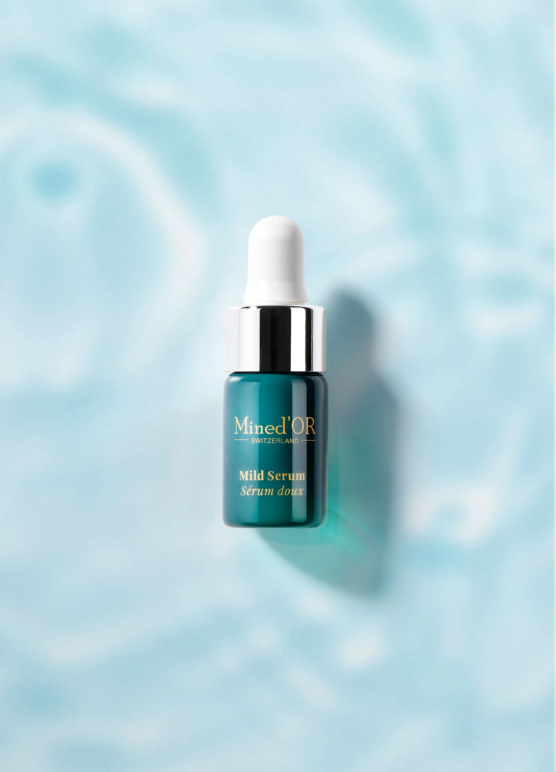 Une bouteille de sérum hydratant de la marque Mined'OR Switzerland, avec un bouchon blanc et glissant, sur un fond bleu clair avec un effet nuageux.