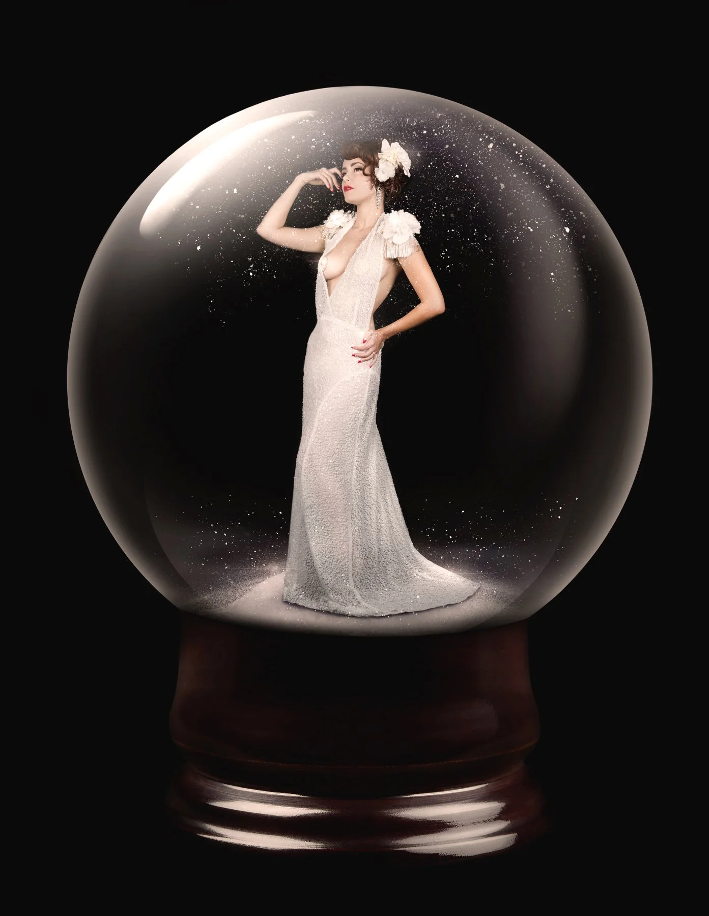 Une femme élégante dans une robe blanche longue et ornée de fleurs, posant sous une boule de verre avec un fond noir.