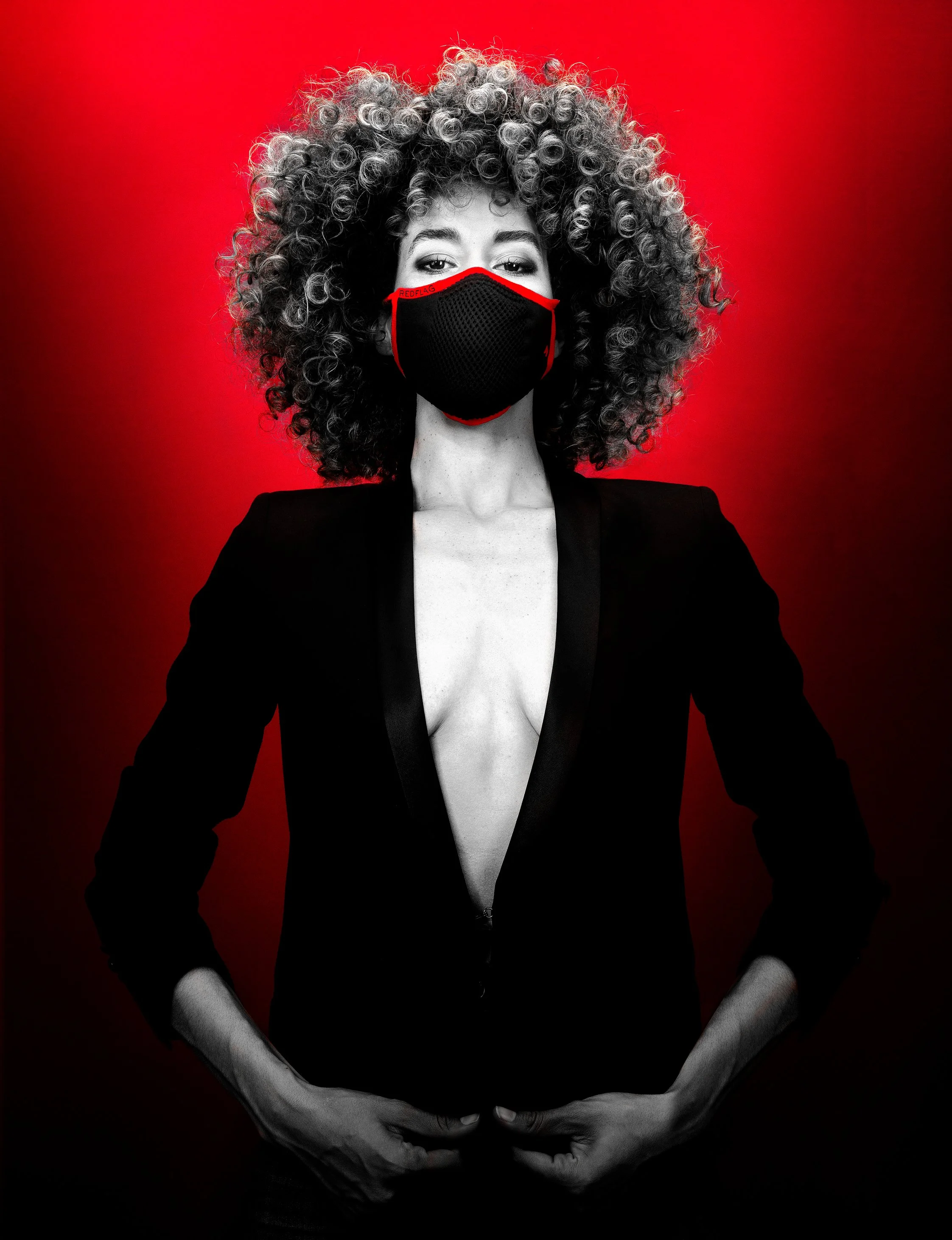 Une femme noire avec de cheveux bouclés porte un masque noir avec une bordure rouge, un manteau noir et se tient contre un fond rouge.