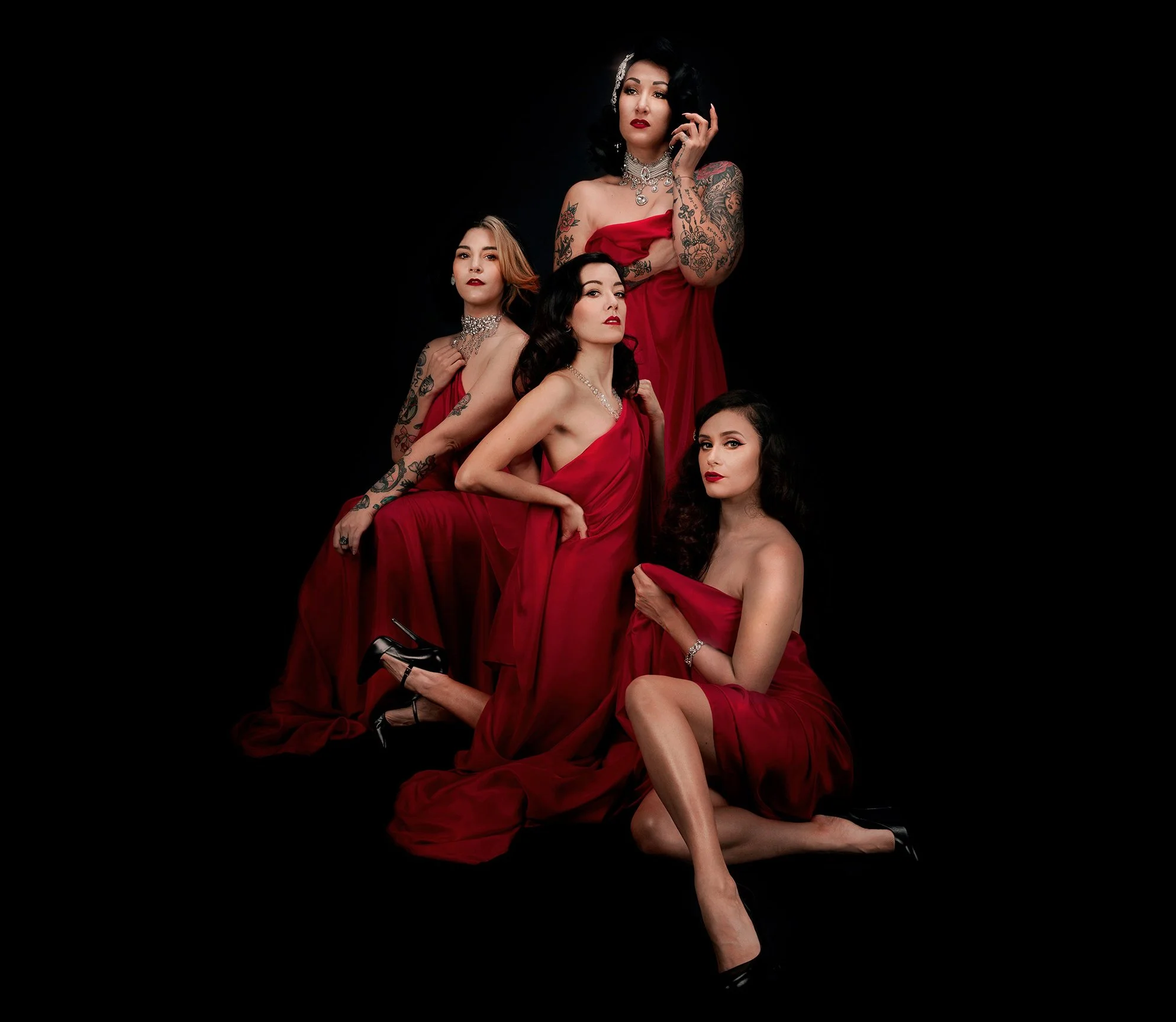 Cinq femmes portant des robes rouges, avec des tatouages, des bijoux et des coiffures vintage, posent contre un fond noir.